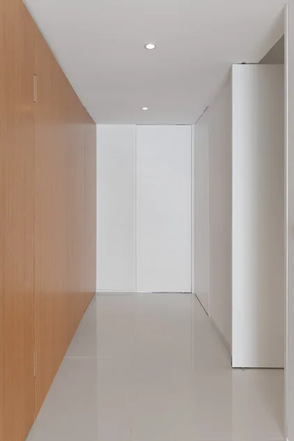A narrow minimalist hallway features floor-to-ceiling wood wall paneling on one side, crisp white walls, and a glossy, reflective floor.