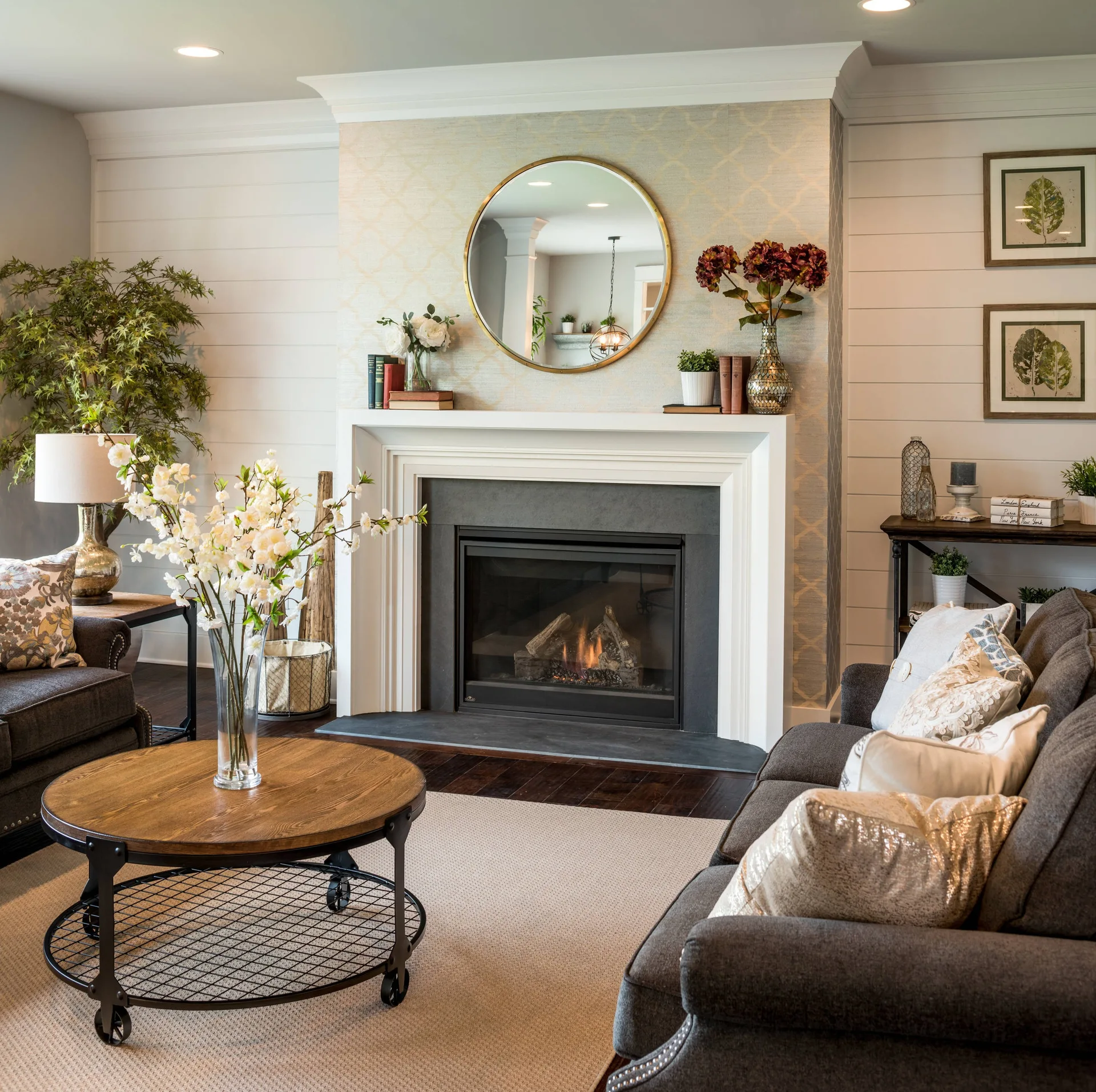 A cozy living room features a fireplace with patterned wallpaper and a white mantel, set against horizontal shiplap walls with a dark gray sofa.