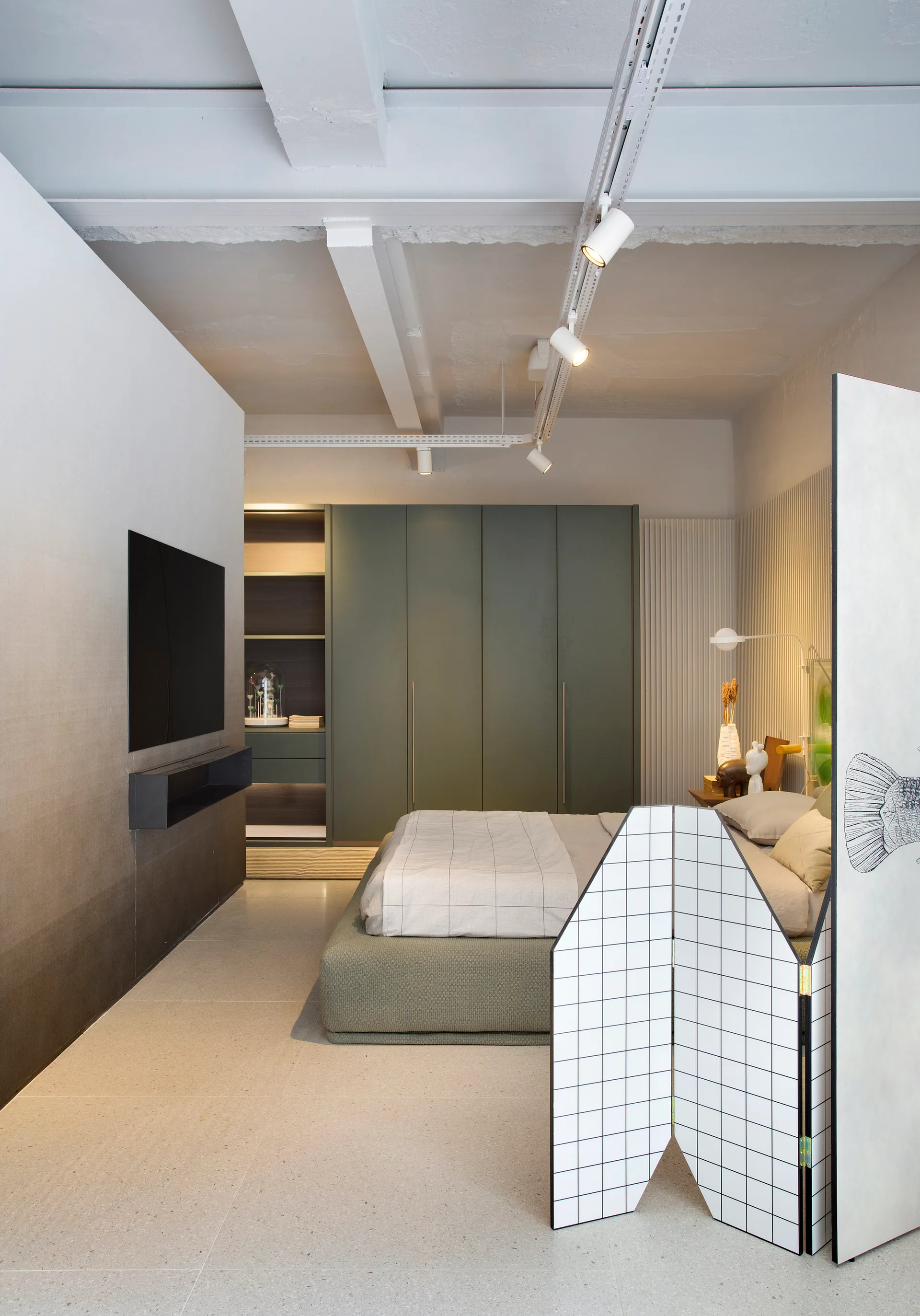 A modern bedroom with a low-profile green bed next to a large sage green wardrobe, with white track lighting on an exposed concrete ceiling.