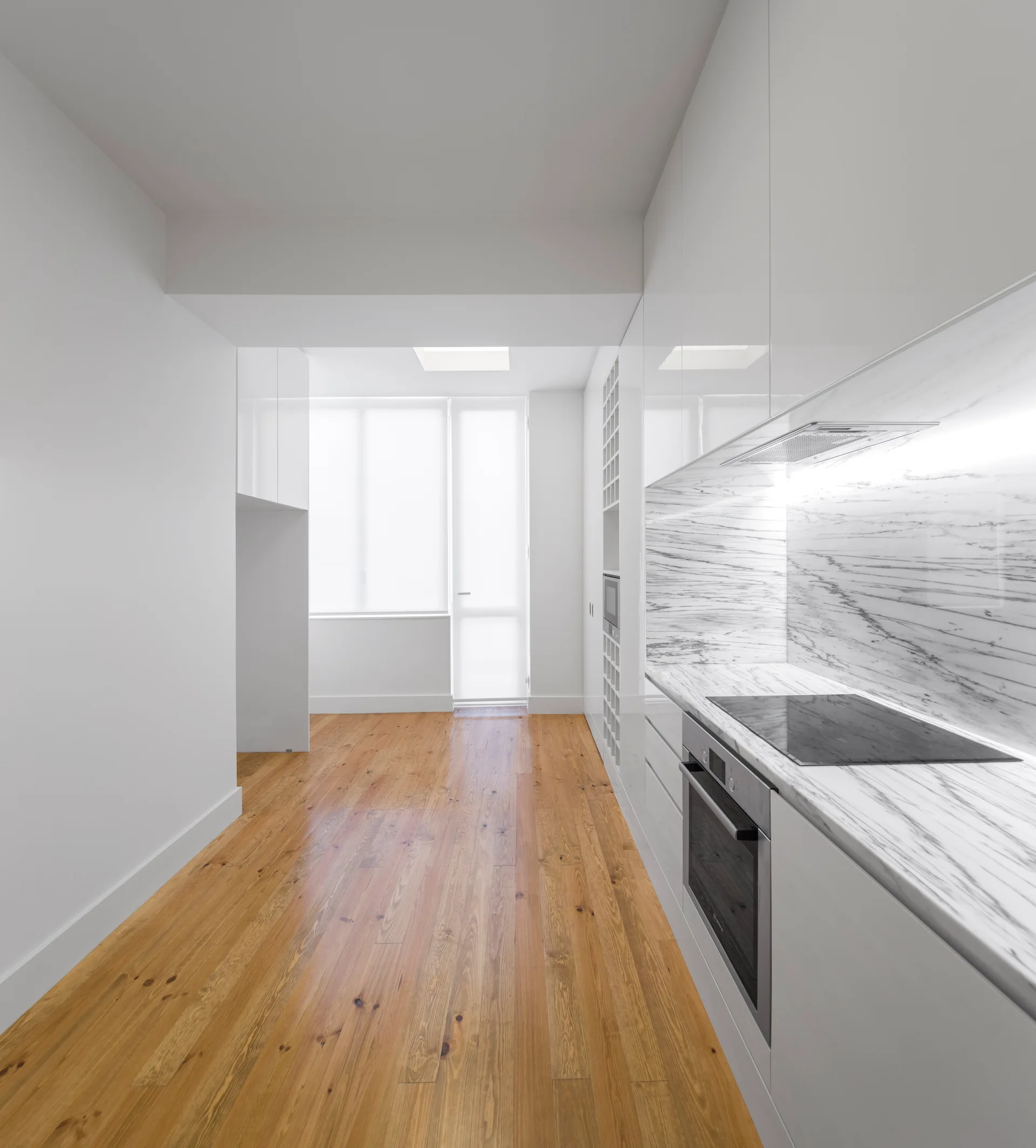 A narrow galley kitchen features glossy white handleless cabinets, a veined marble countertop and backsplash, and light wood plank flooring.