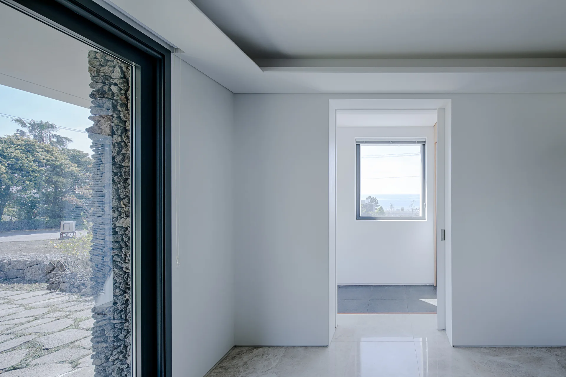 A minimalist white hallway with glossy marble floors, a recessed ceiling, and an open doorway leading to a room with a square window.