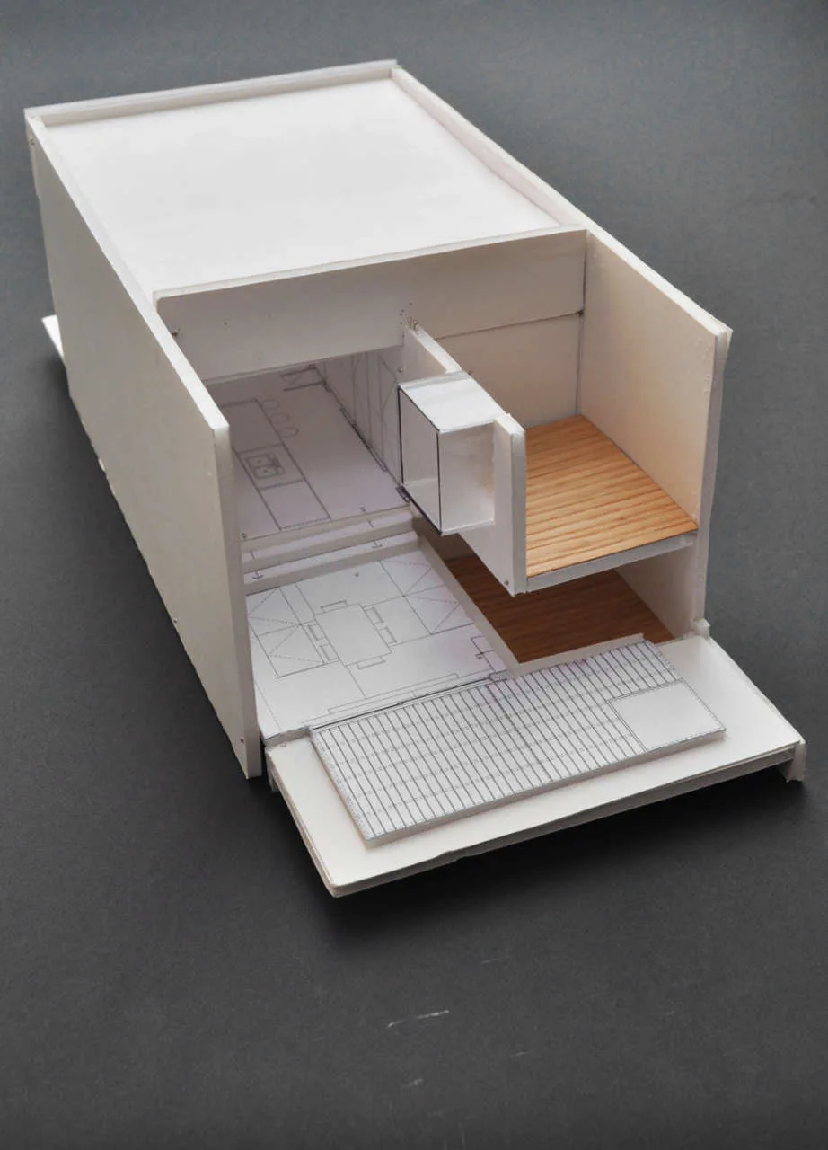 A white cardboard architectural scale model showing a split-level interior layout with wooden floor sections and printed floor plans.