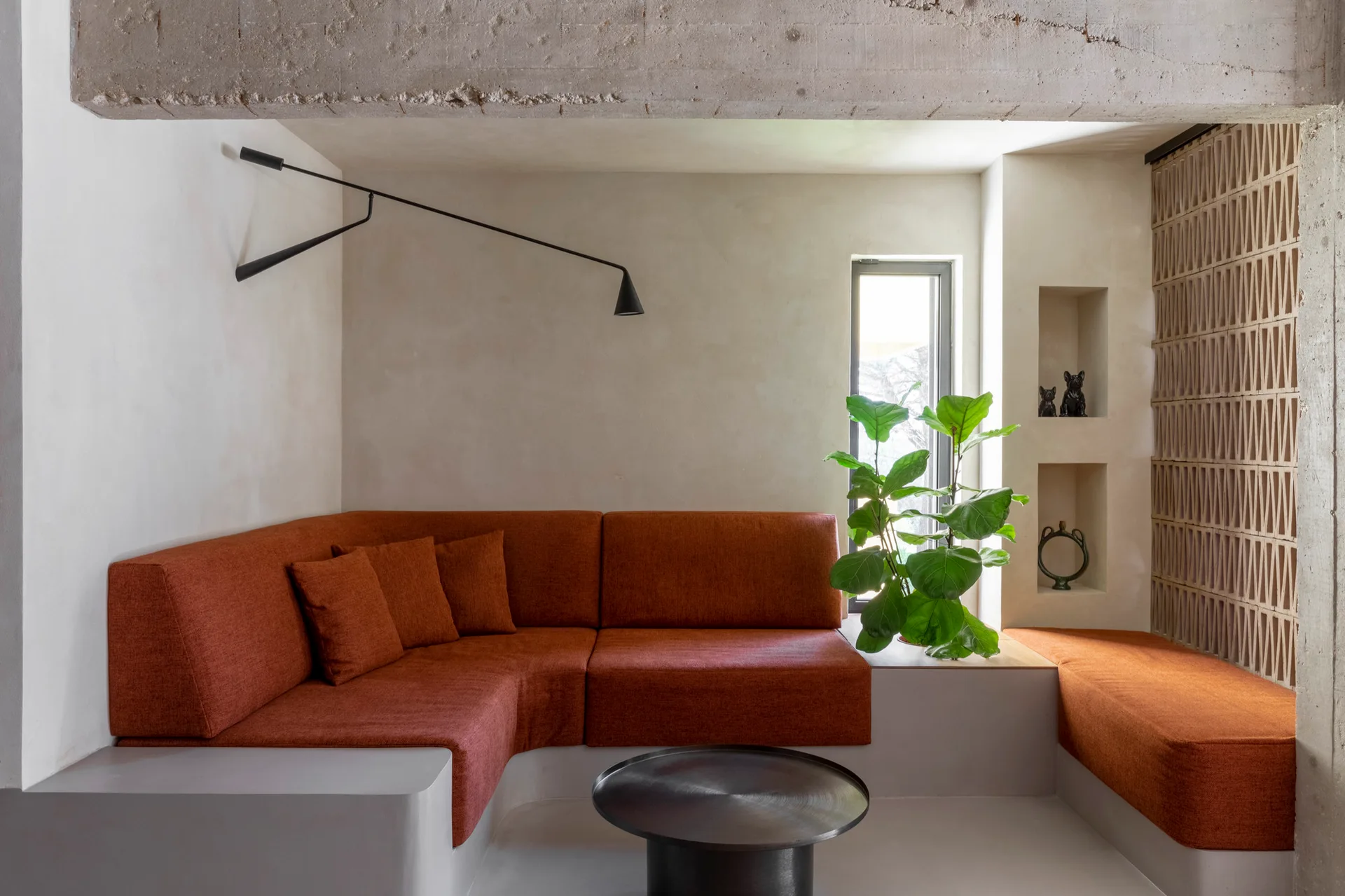 A built-in rust orange sectional sofa on a grey concrete base in a minimalist room with a raw concrete beam, wall niches, and a breeze block screen.