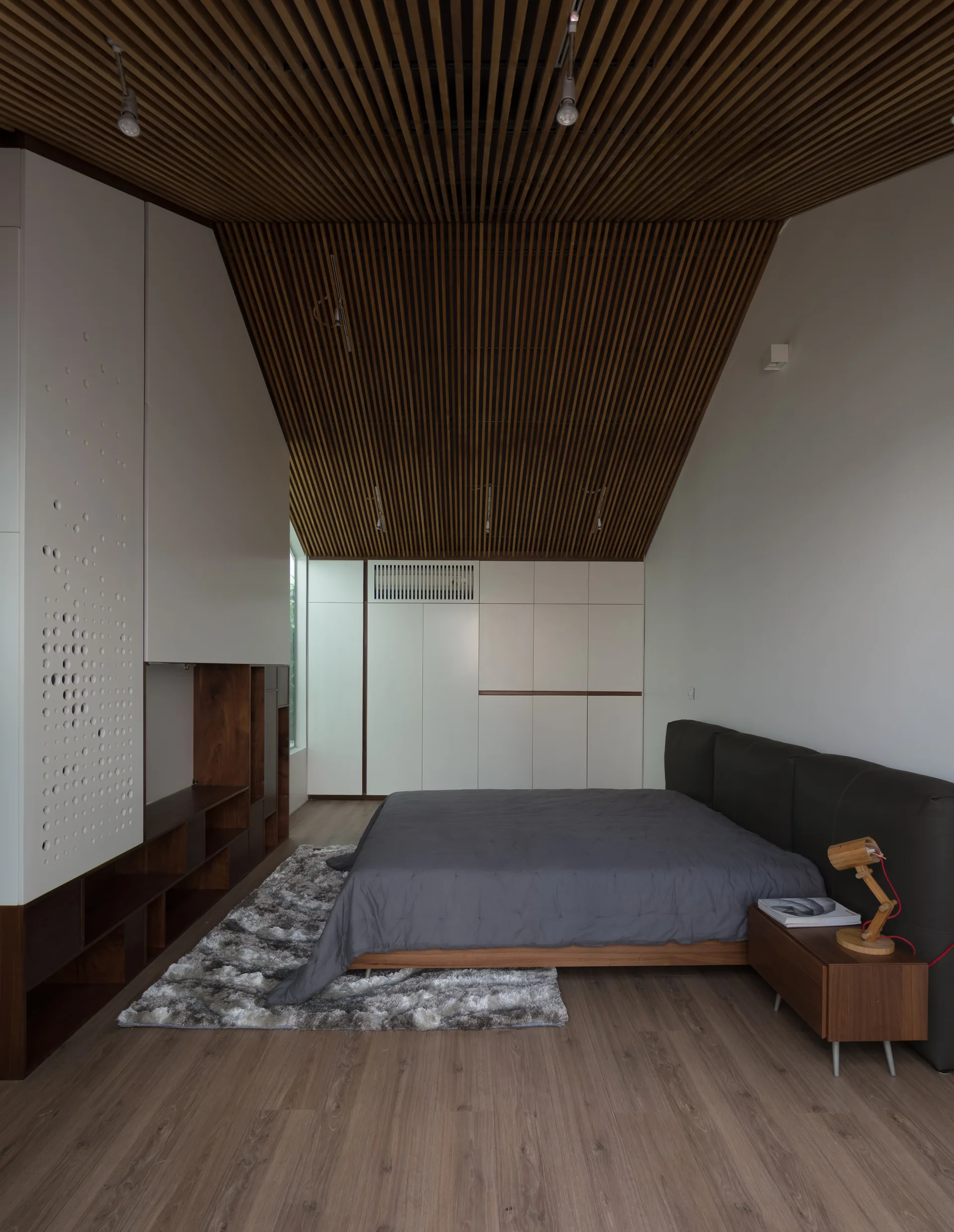 A minimalist bedroom featuring a sloped wood slat ceiling, built-in white cabinets, a grey bed, and a textured area rug.
