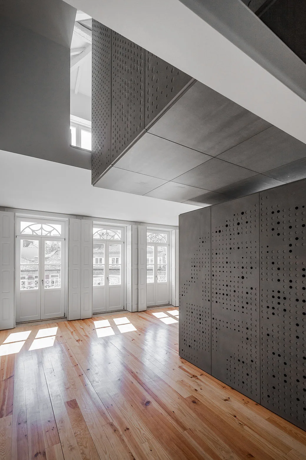 A bright room with natural wood flooring features white French doors and a massive gray perforated architectural partition extending to the upper level.