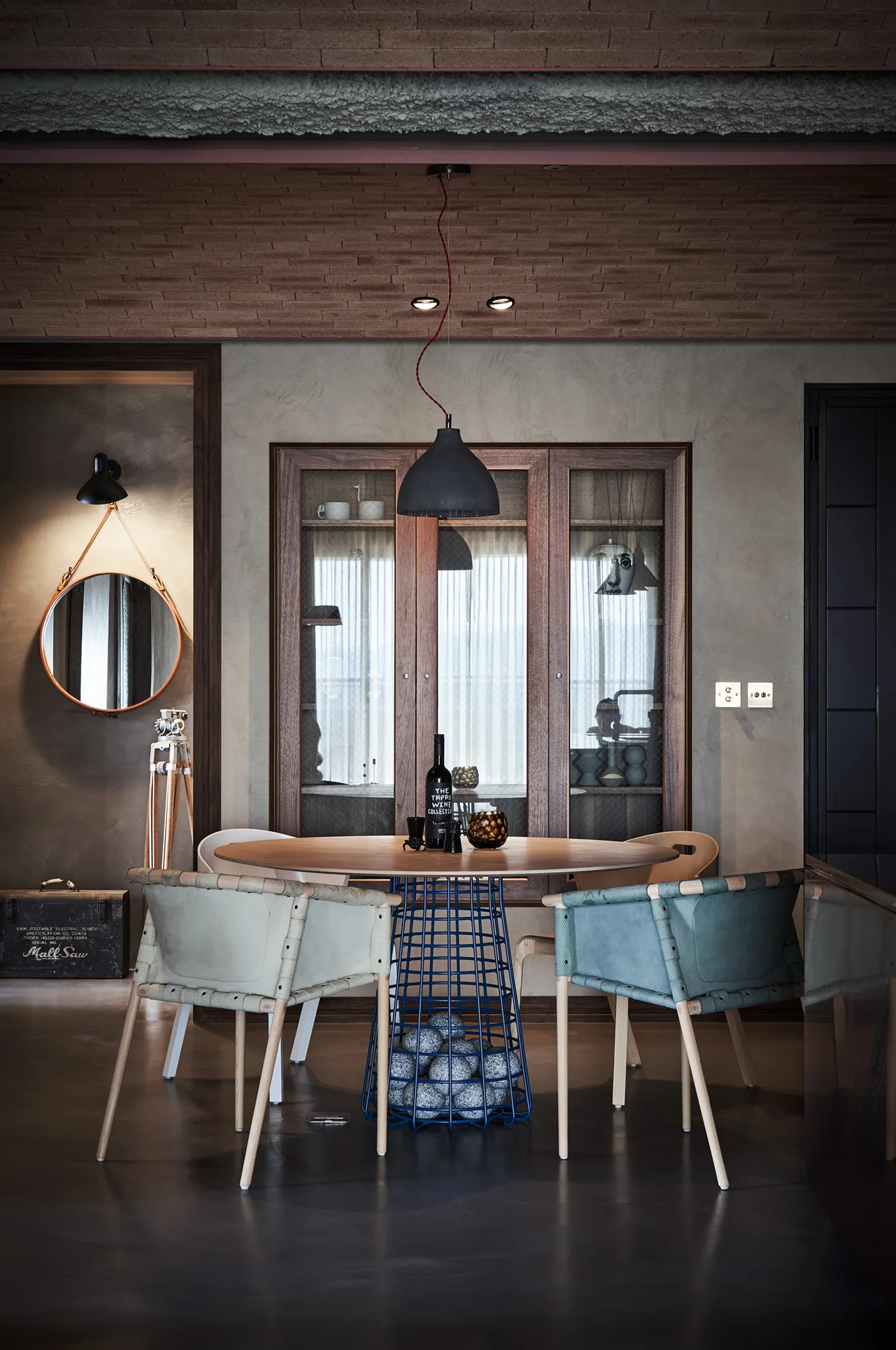 A modern industrial dining room features a round wooden table with a blue wire pedestal base, leather sling chairs, and an exposed brick ceiling.