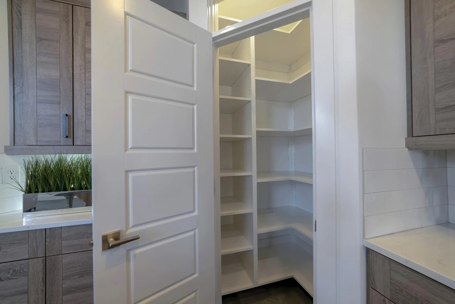 An open white paneled door reveals a walk-in pantry with white tiered shelving, framed by grey wood-grain kitchen cabinets.