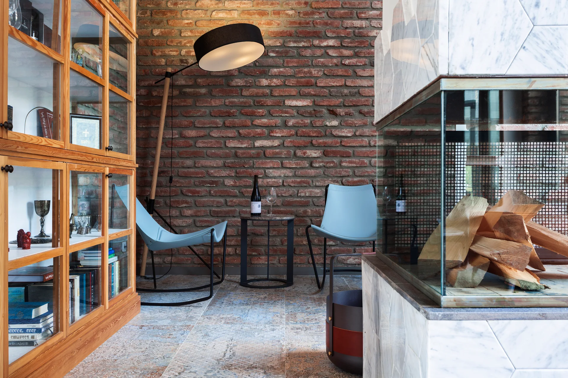 A cozy sitting nook features two light blue leather chairs against an exposed brick wall next to a wooden display cabinet and a marble fireplace.