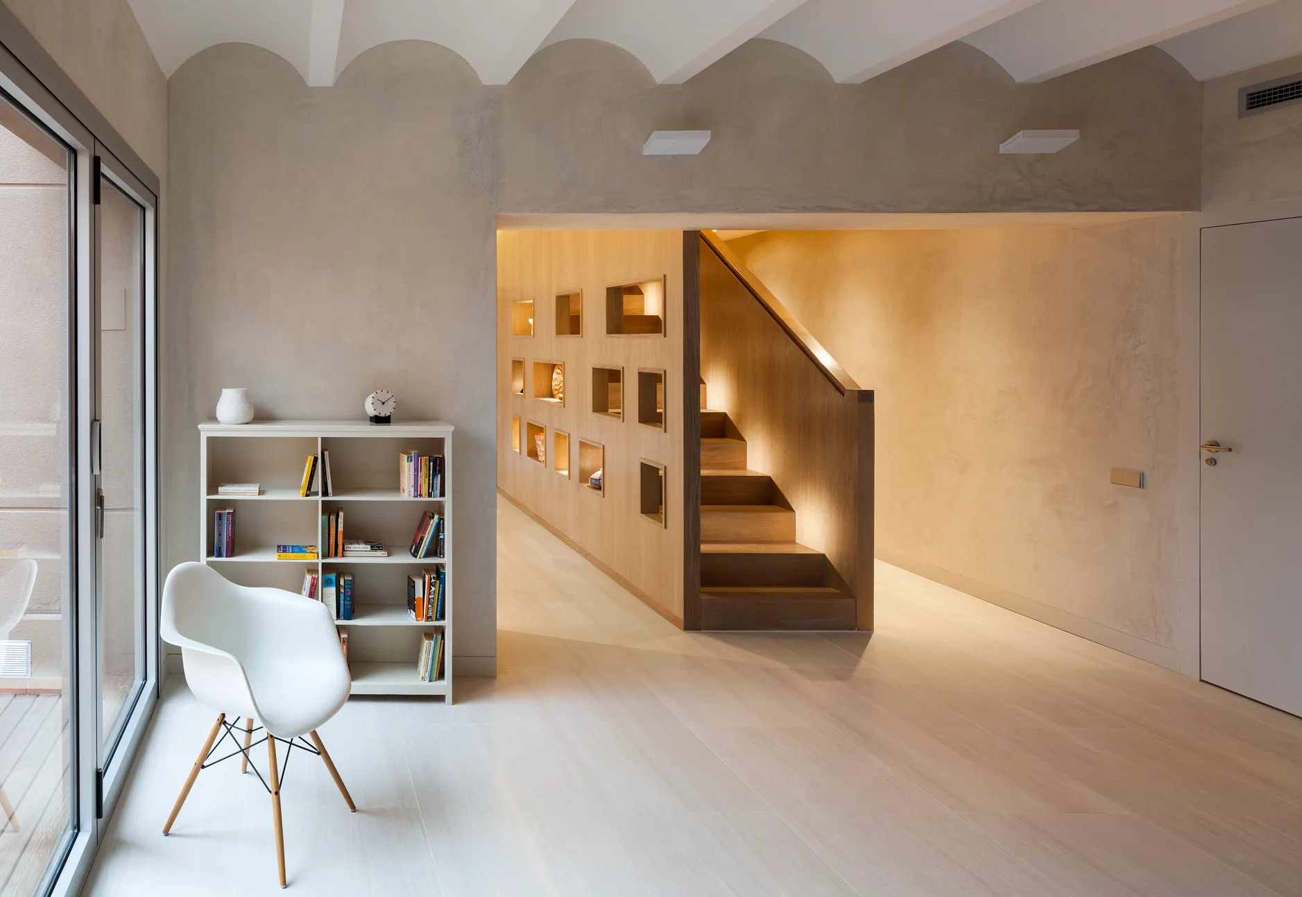 A wooden staircase with illuminated built-in wall niches stands next to plaster walls, an arched ceiling, and a white chair.