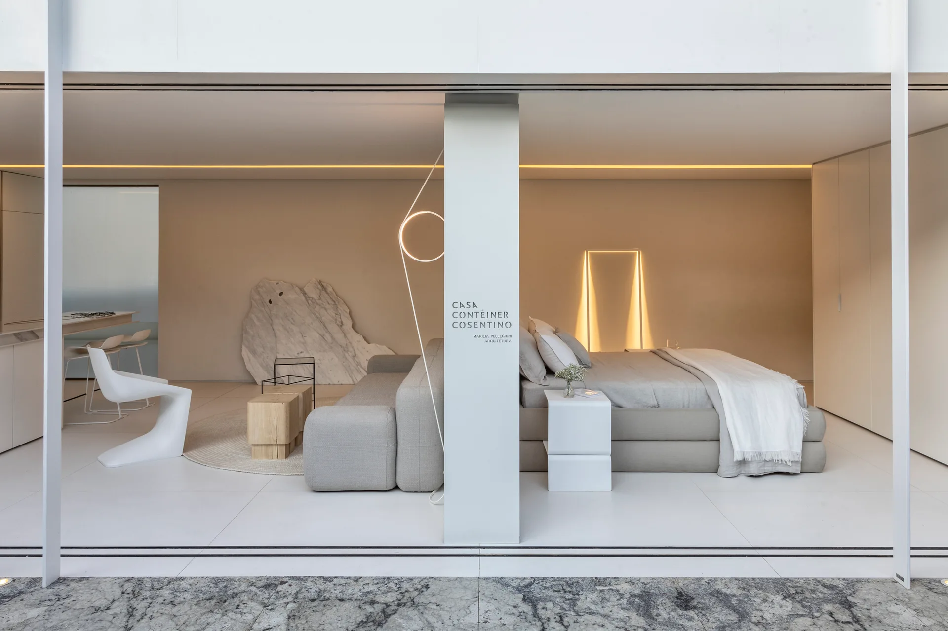 A minimalist open-plan studio featuring a grey upholstered bed and modular sofa divided by a central white column under warm cove lighting.