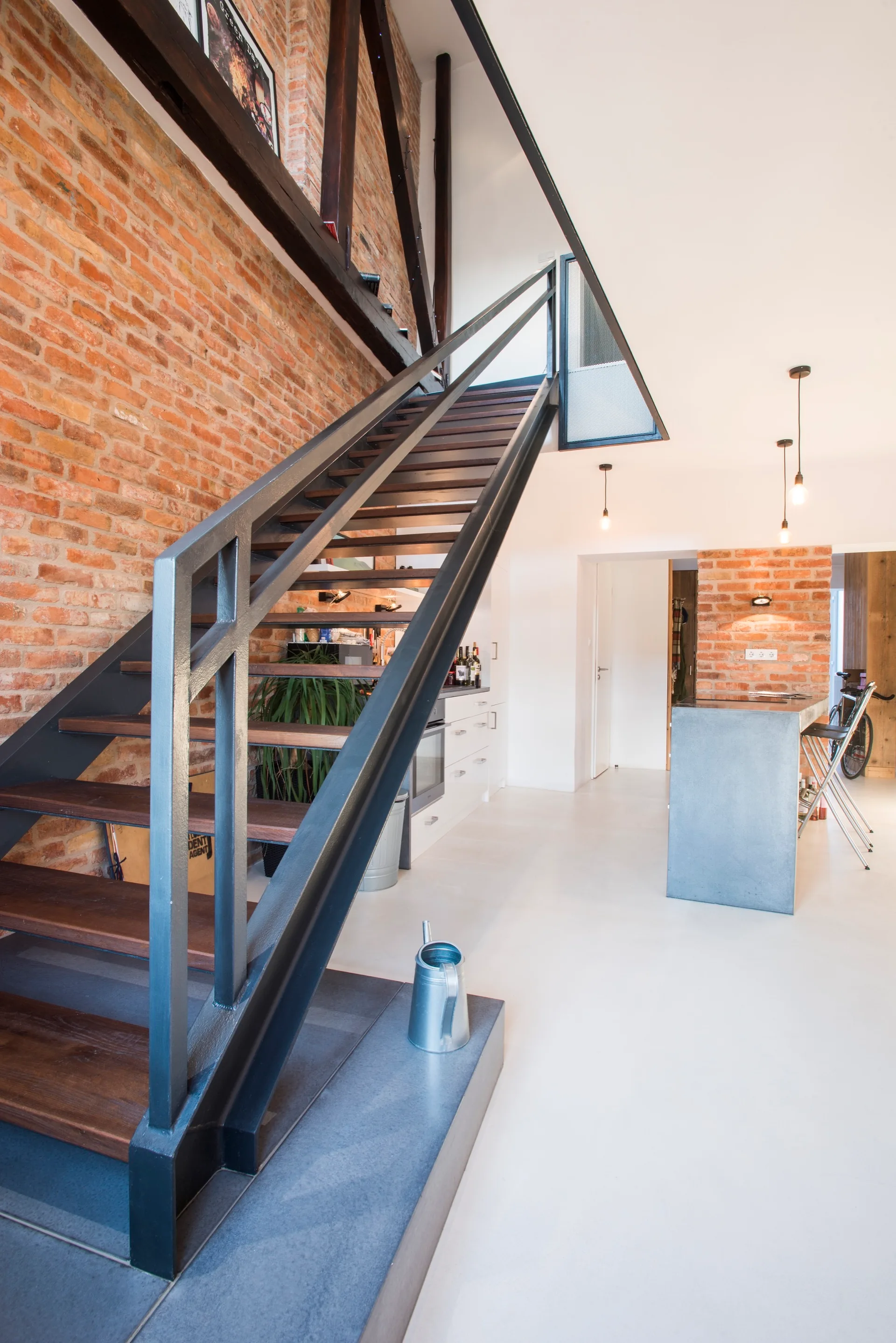 An open-riser black metal staircase with wood treads stands against a rustic exposed brick wall next to a modern kitchen area with light flooring.