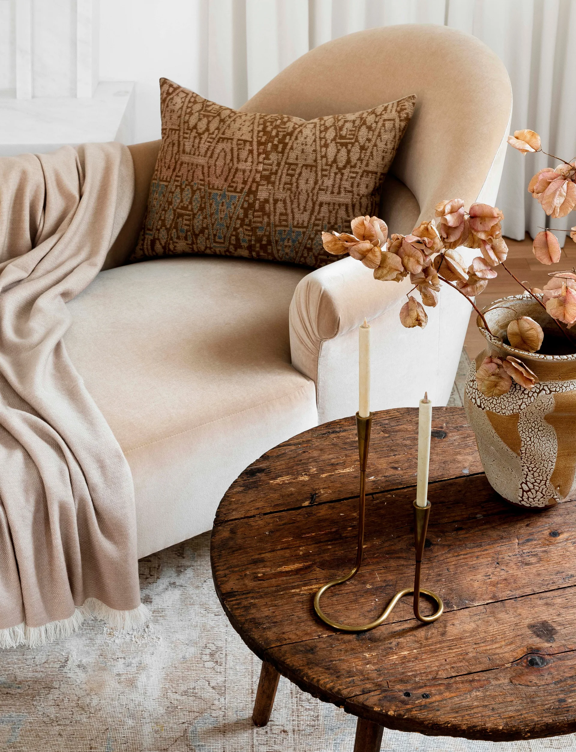 A curved beige chaise lounge with a patterned pillow and fringed throw sits next to a round rustic wooden side table decorated with taper candles.