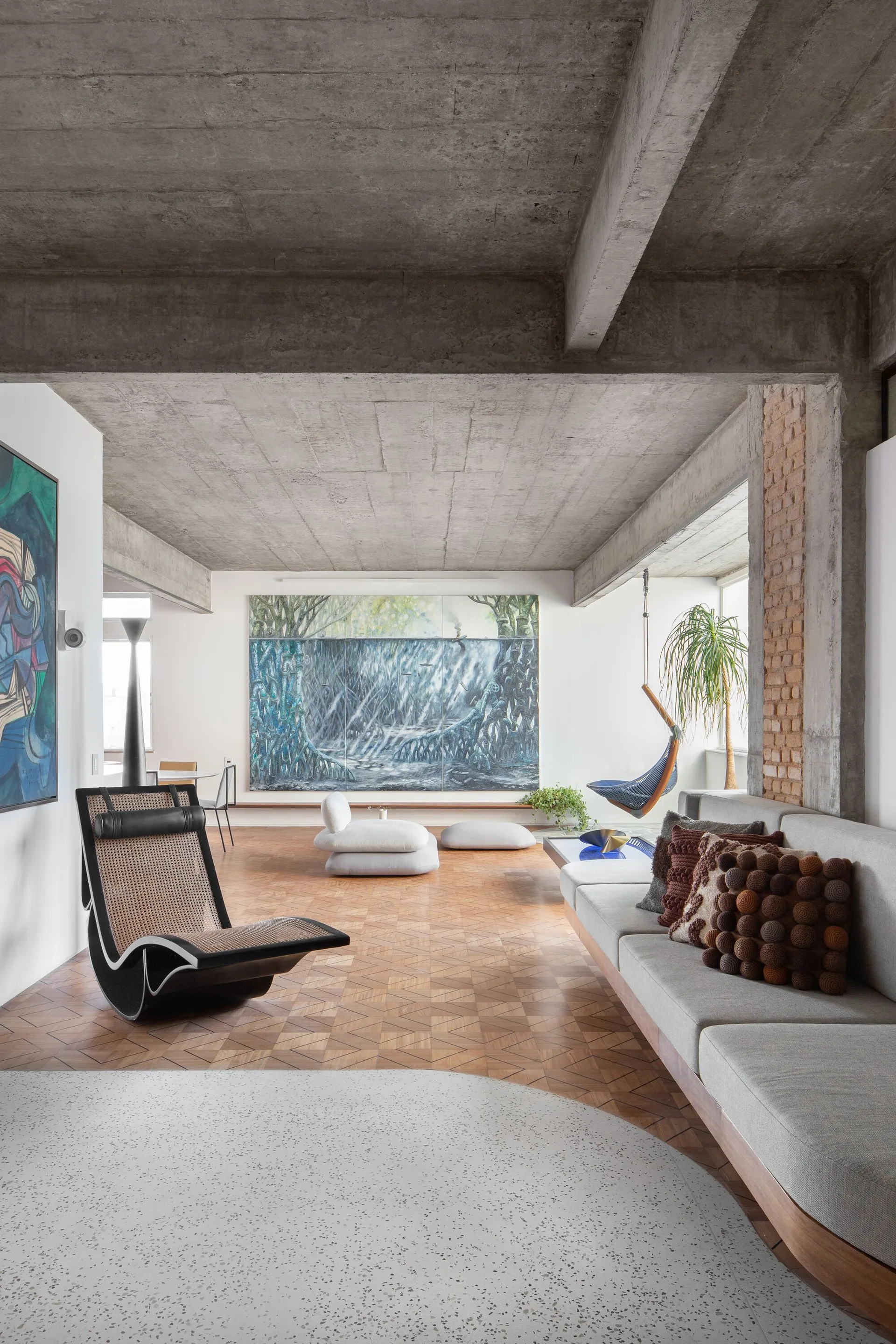 A spacious modern living room features an exposed concrete ceiling, a grey sofa with textured pillows, and patterned parquet wood floors.