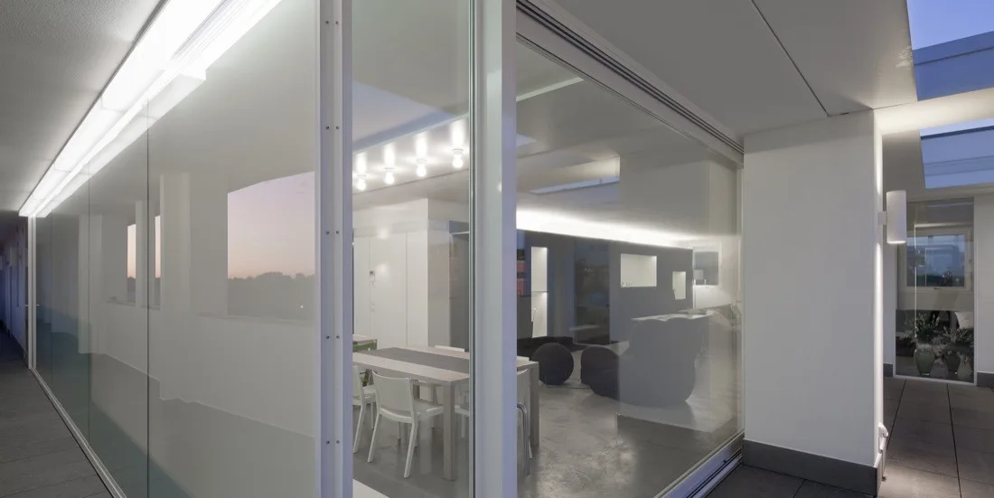 A modern apartment dining and living room seen through a large floor-to-ceiling sliding glass wall next to an outdoor stone terrace.