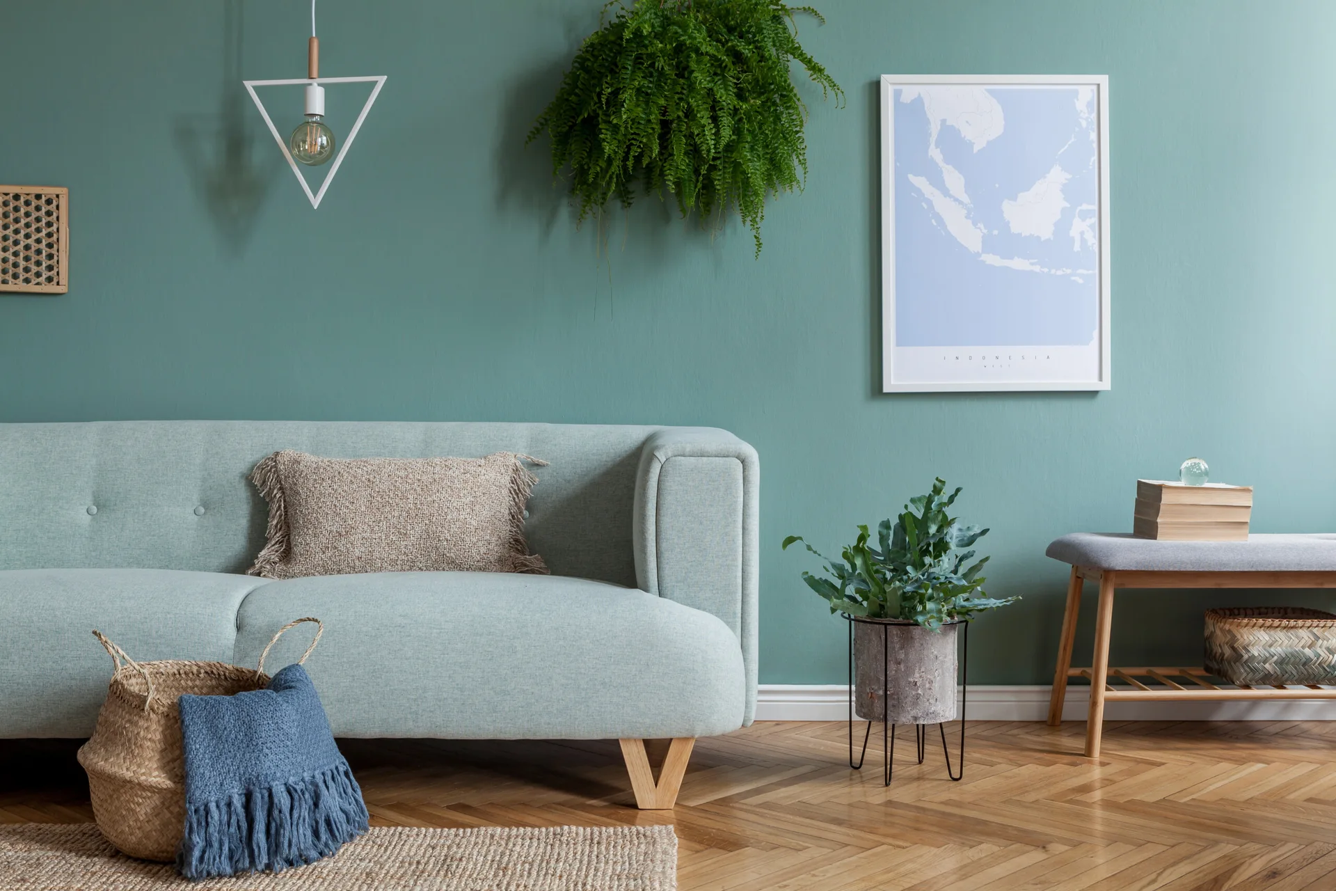 A light blue sofa sits against a solid teal wall in a living room featuring herringbone wood floors, a hanging plant, and a framed map.