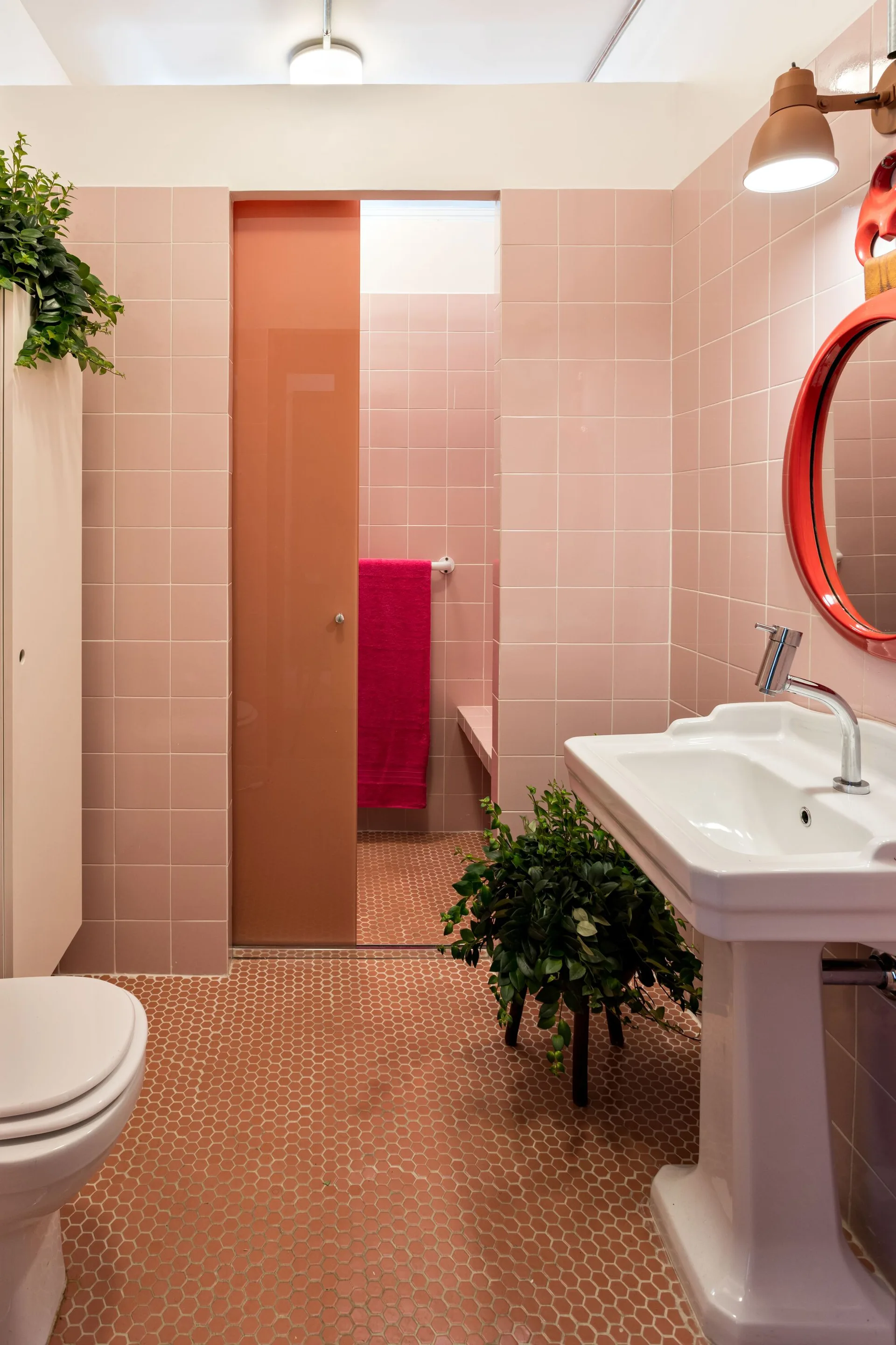 A bathroom features pink square-tiled walls, an orange hexagonal tile floor, a white pedestal sink, and a round red-framed mirror.