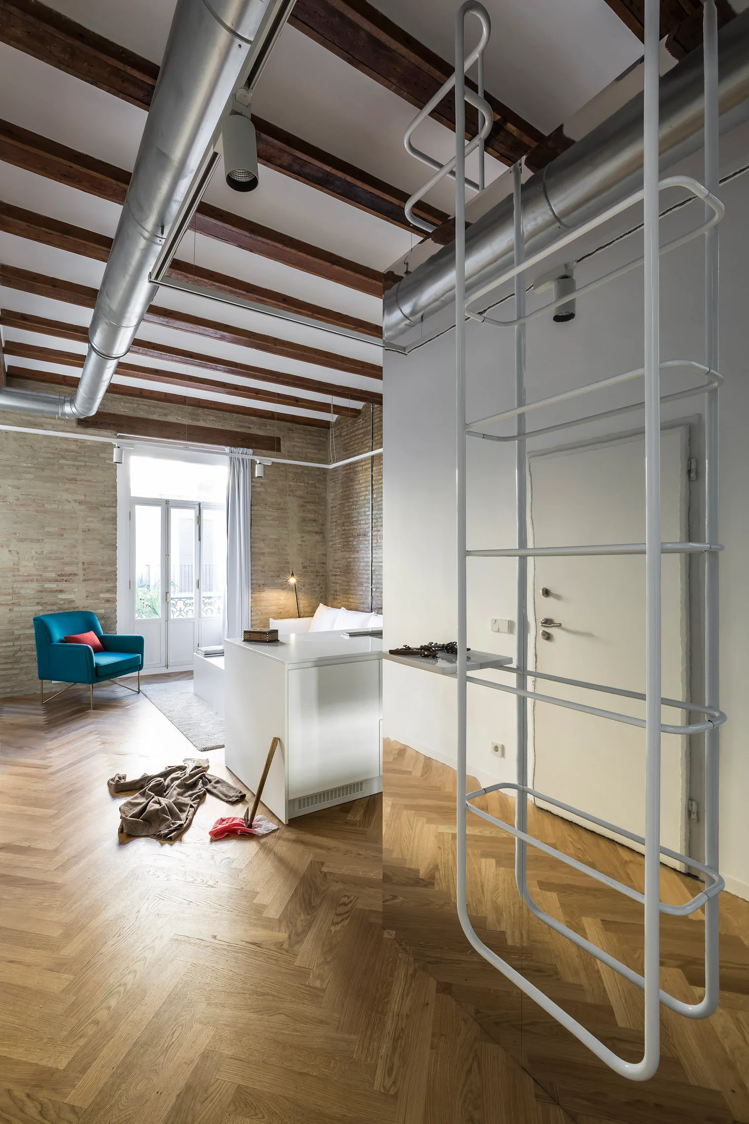 A modern loft apartment featuring herringbone wood floors, exposed brick walls, dark ceiling beams, industrial metal ductwork, and a white metal ladder.