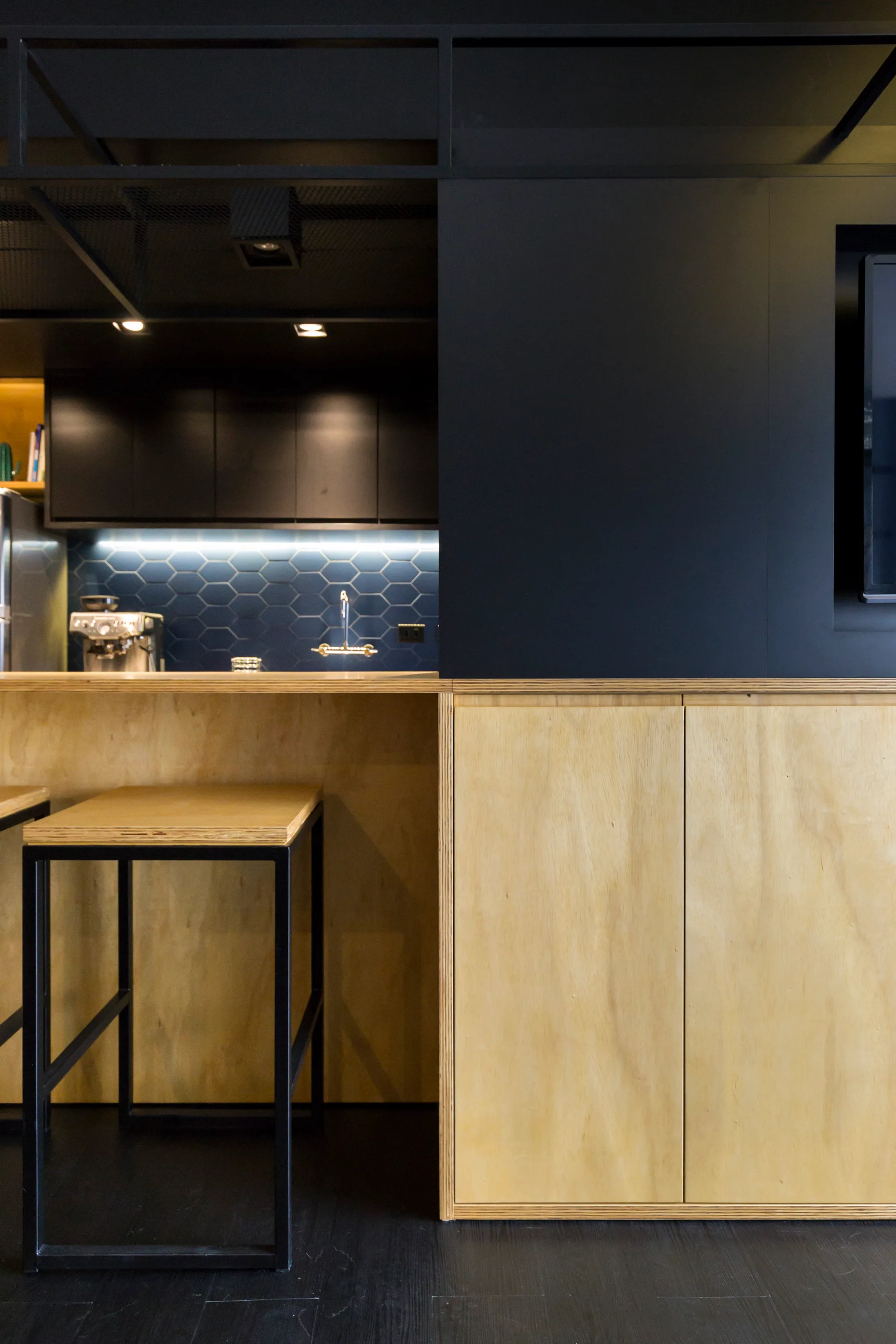 A modern kitchen featuring a plywood island with bar stools, matte black upper cabinets, and a blue hexagonal tile backsplash.