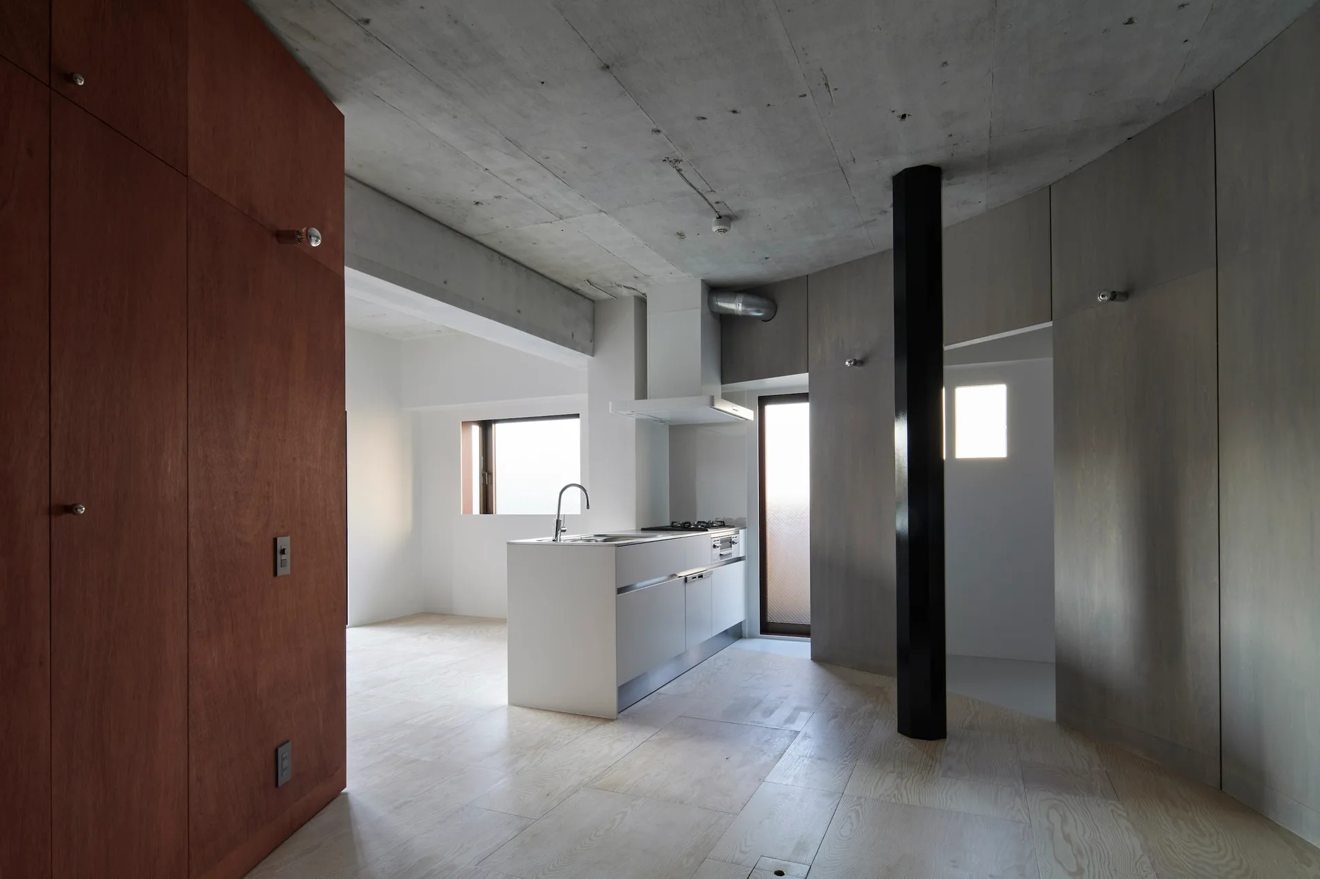 A minimalist kitchen features a white island, a raw concrete ceiling, a black structural column, and warm wood paneled walls.