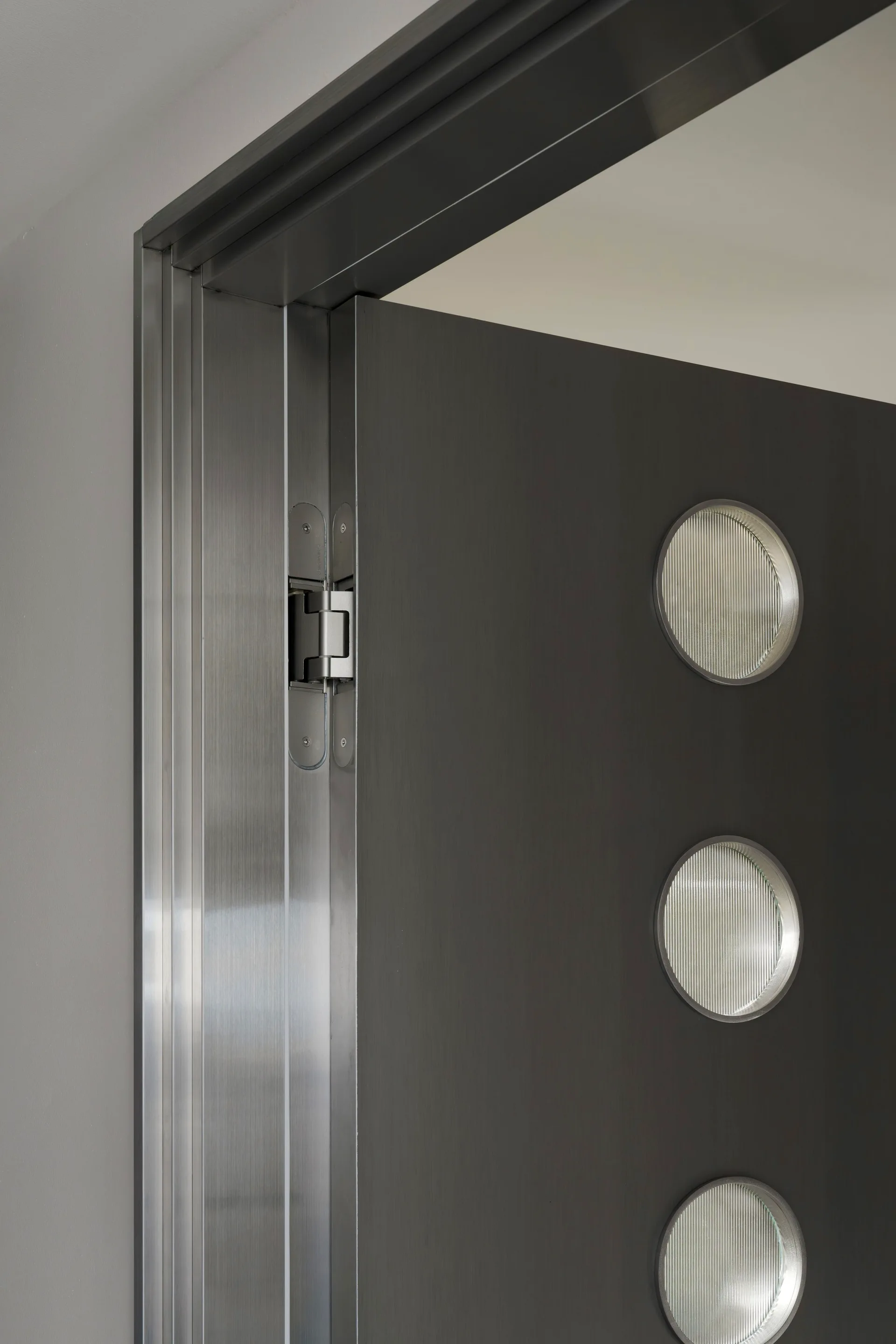 A close-up of a dark grey door with a brushed metal frame, a concealed hinge, and three circular ribbed glass windows.