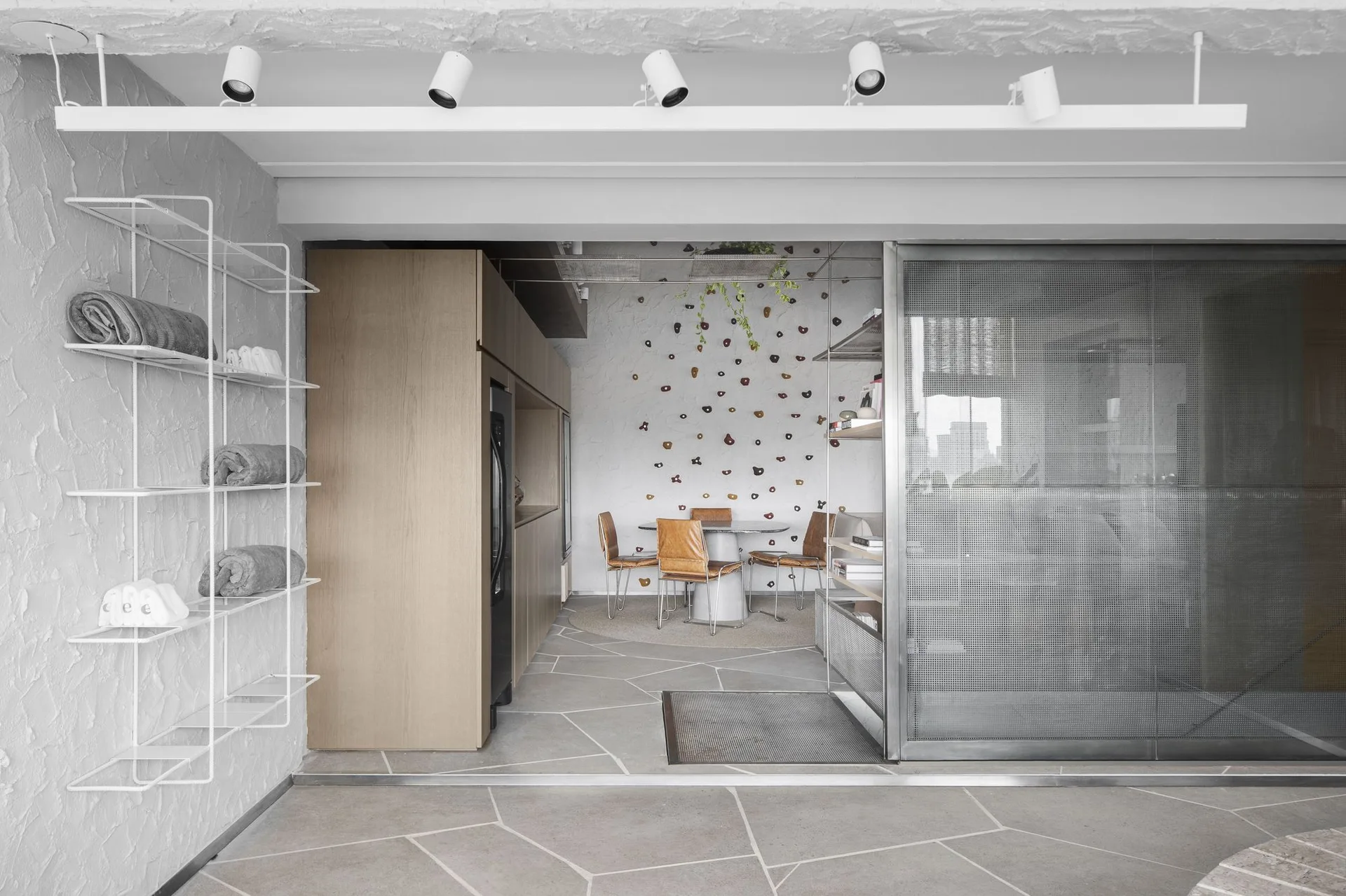 A modern open-plan dining area features a climbing wall with colorful holds, tan leather chairs, wood cabinetry, and a metal mesh partition.
