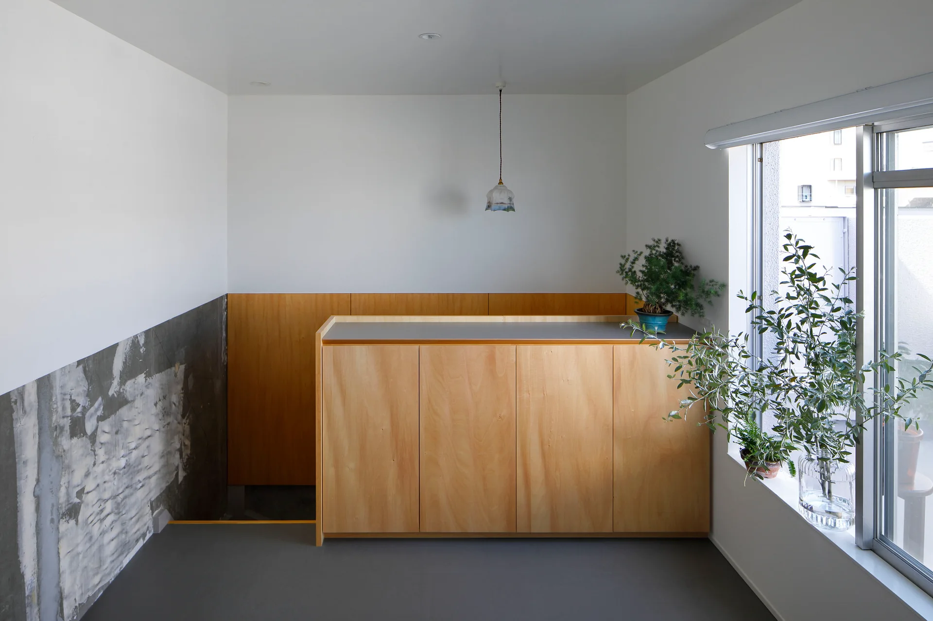 A minimalist interior featuring a custom wood cabinet, a raw textured concrete wall, grey flooring, and a window with green plants.