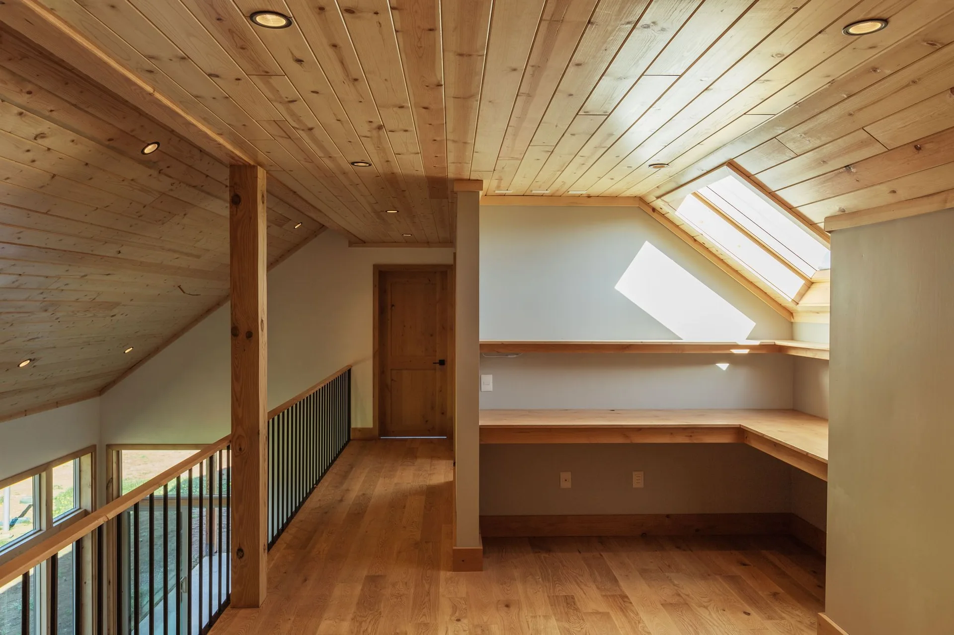 A loft landing hallway features a built-in L-shaped wooden desk under a sloped ceiling with a skylight, wood-paneled ceiling, and black metal railing.