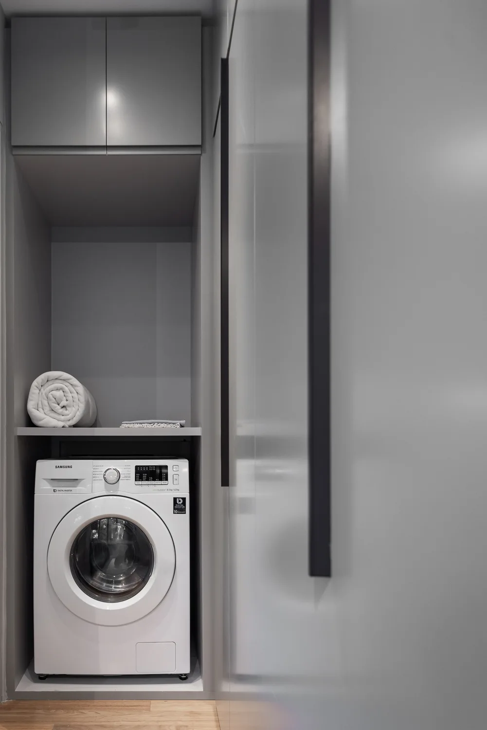 A white front-loading washing machine is integrated into a matte grey built-in cabinet unit with an open storage shelf.