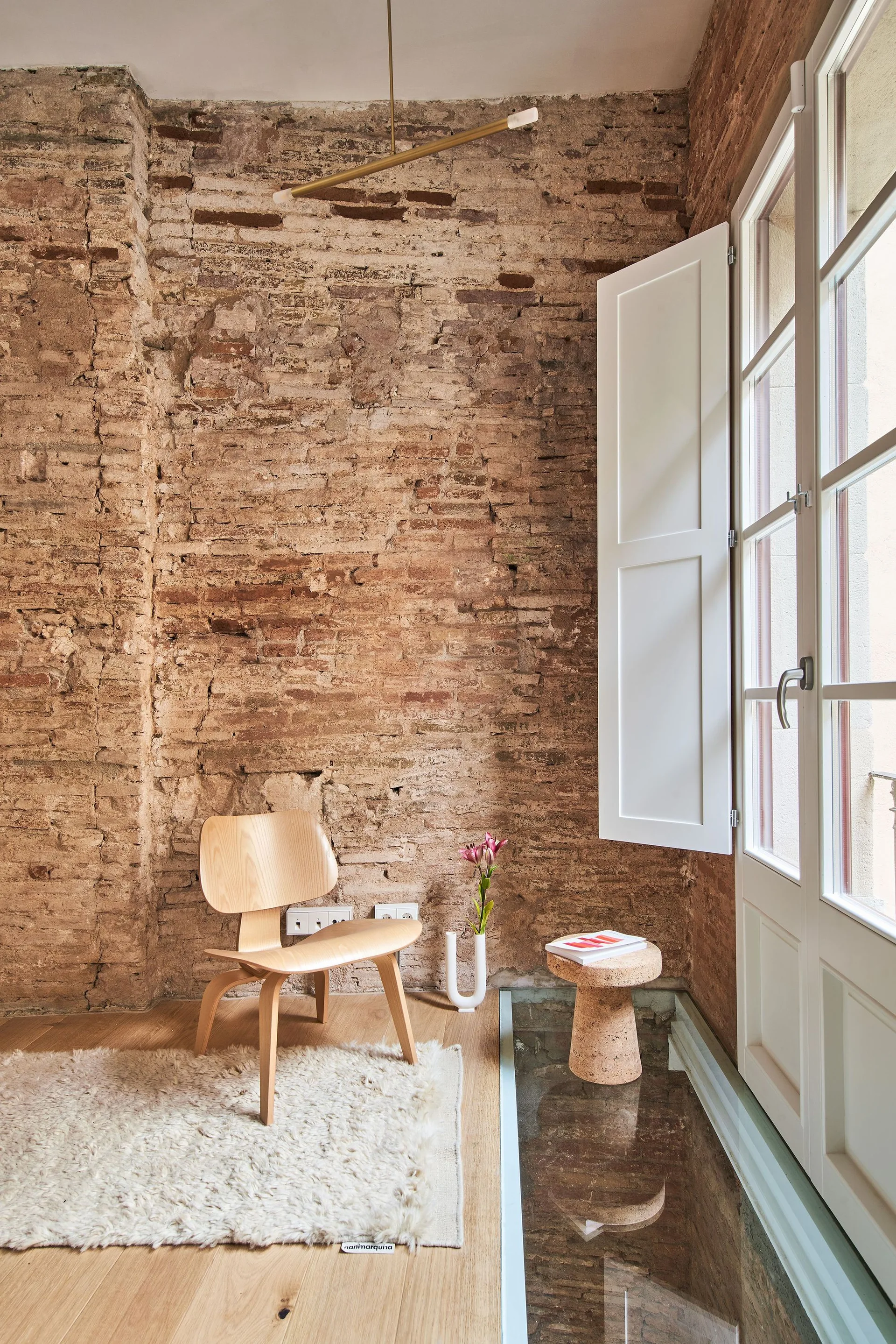 A molded plywood chair sits on a shaggy rug next to an exposed brick wall, a cork side table, and an inset glass floor section.
