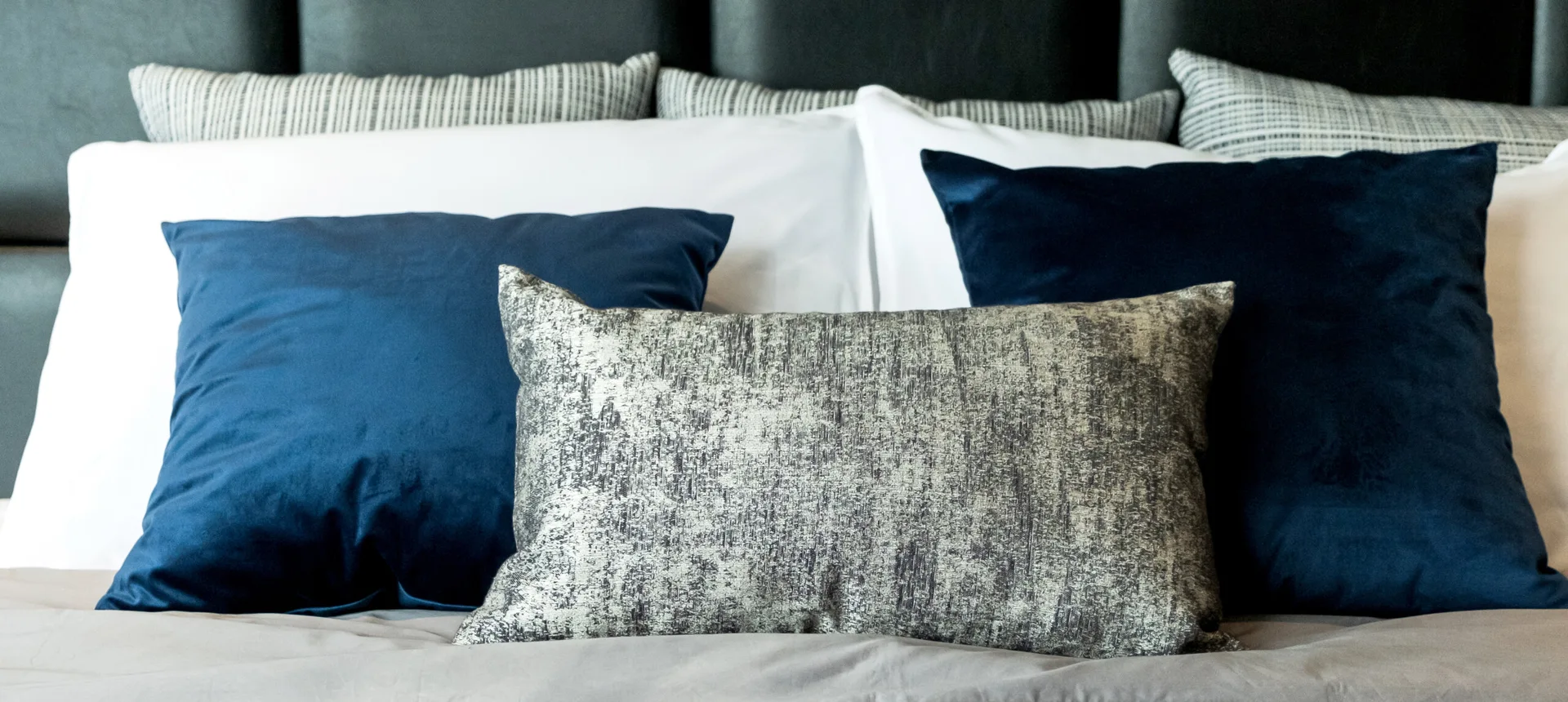 A close-up of layered bed bedding with white shams, navy blue velvet pillows, and a textured silver lumbar pillow against a dark headboard.