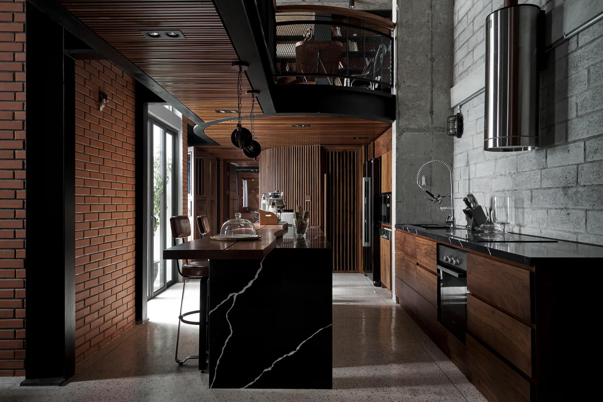 An industrial kitchen features a black veined stone island with stools, dark wood cabinetry, a slatted ceiling, and exposed brick walls.