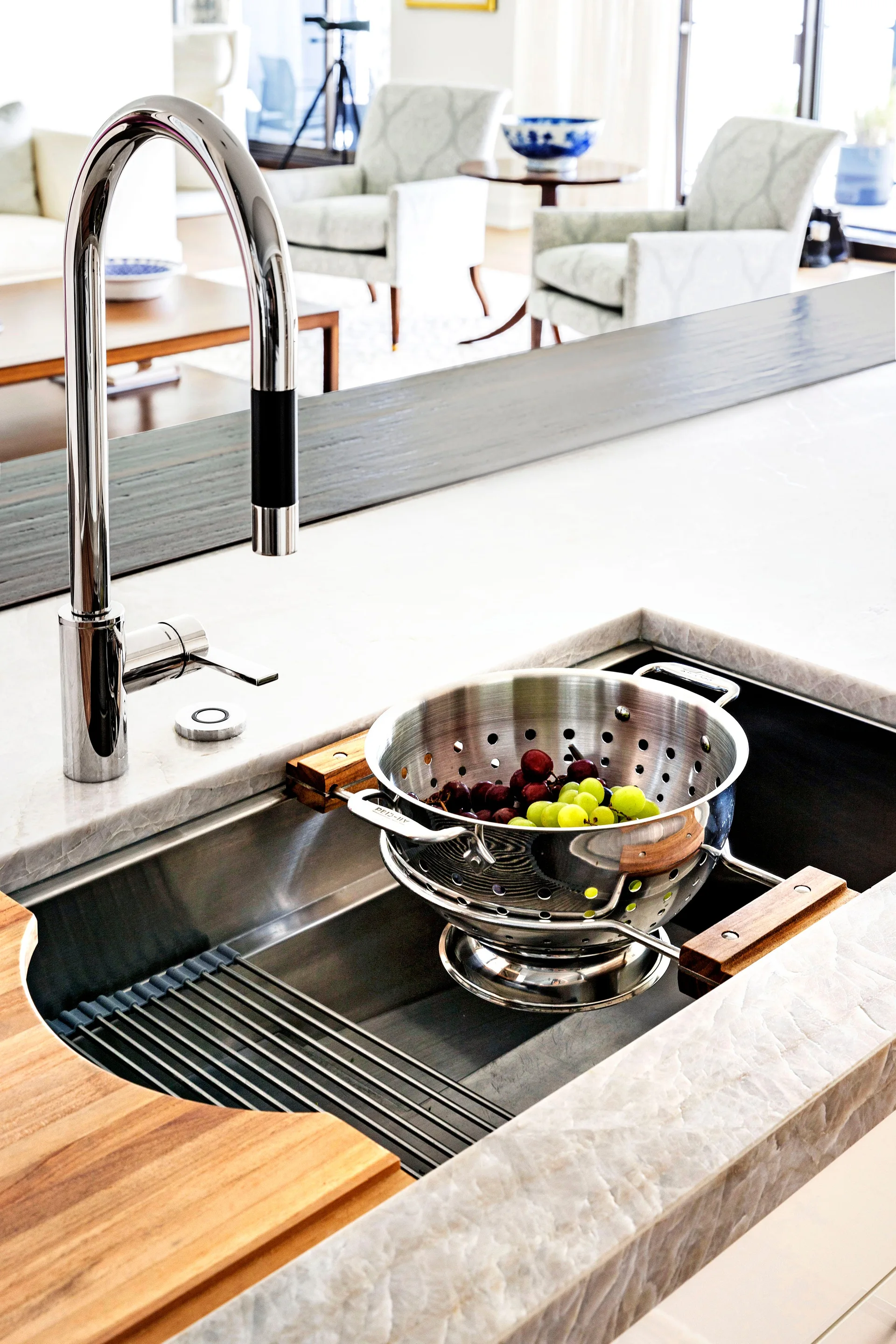A stainless steel undermount kitchen sink set in a light veined countertop with a high-arc faucet and a colander of grapes.