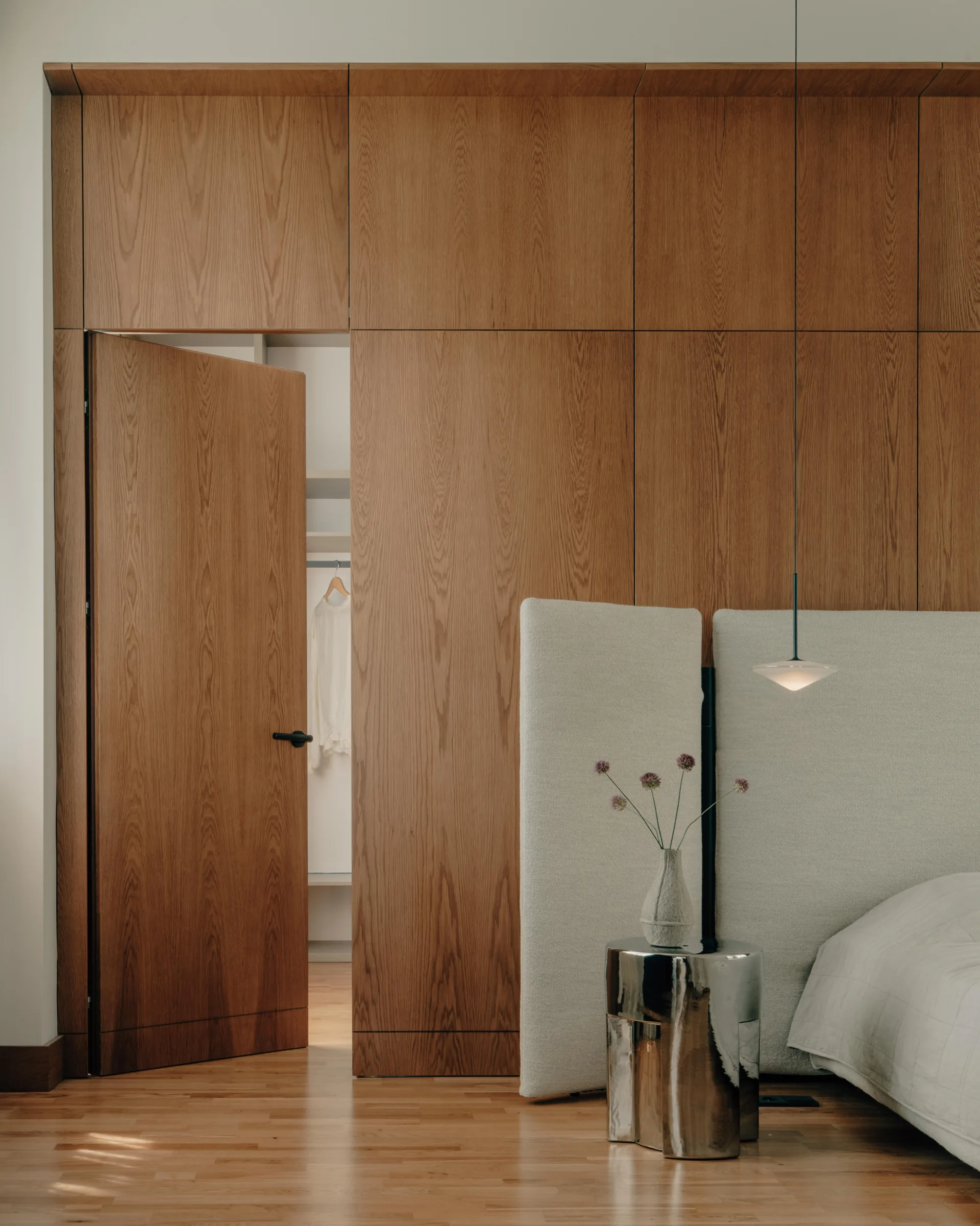 A bedroom featuring a seamless wood-paneled closet wall with a slightly open door, next to a tall cream headboard and a silver metallic side table.