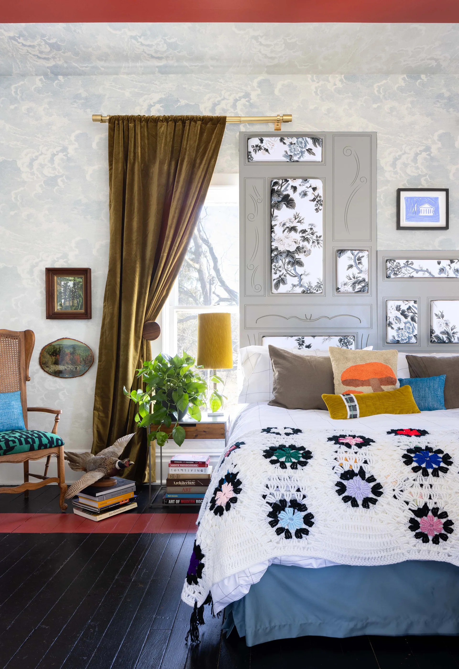 A bedroom features blue cloud-print wallpaper, a grey custom headboard with floral fabric panels, and a colorful crocheted quilt on the bed.