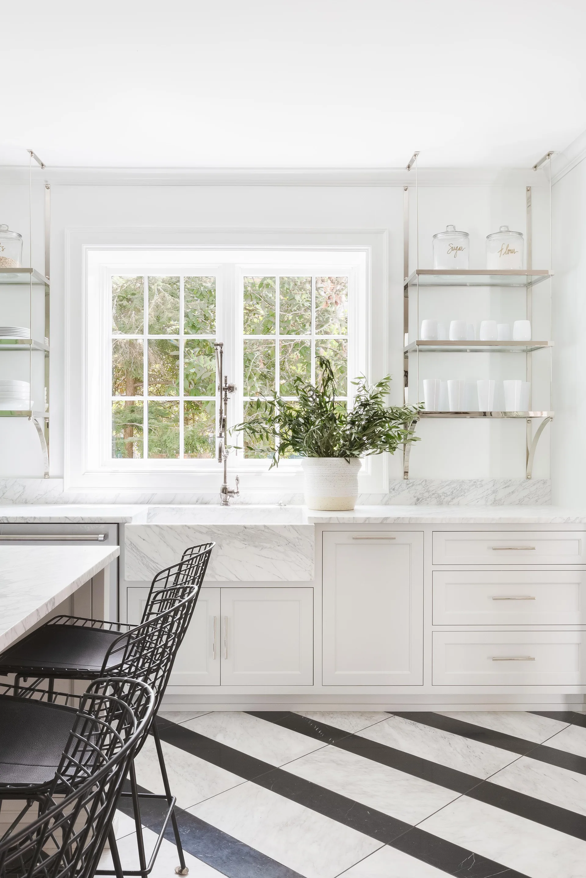 A bright white kitchen with a marble farmhouse sink, white cabinets, polished metal open shelves, and diagonal black and white tiled floors.