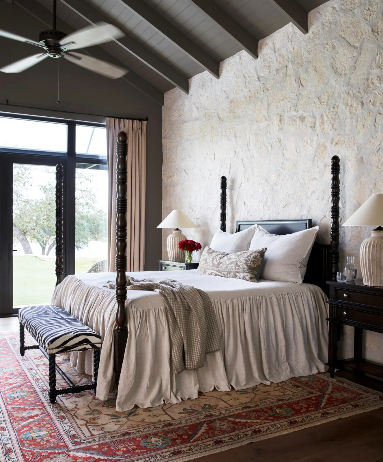 A bedroom features a dark wood four-poster bed set against a white natural stone wall, underneath a dark-painted vaulted ceiling.