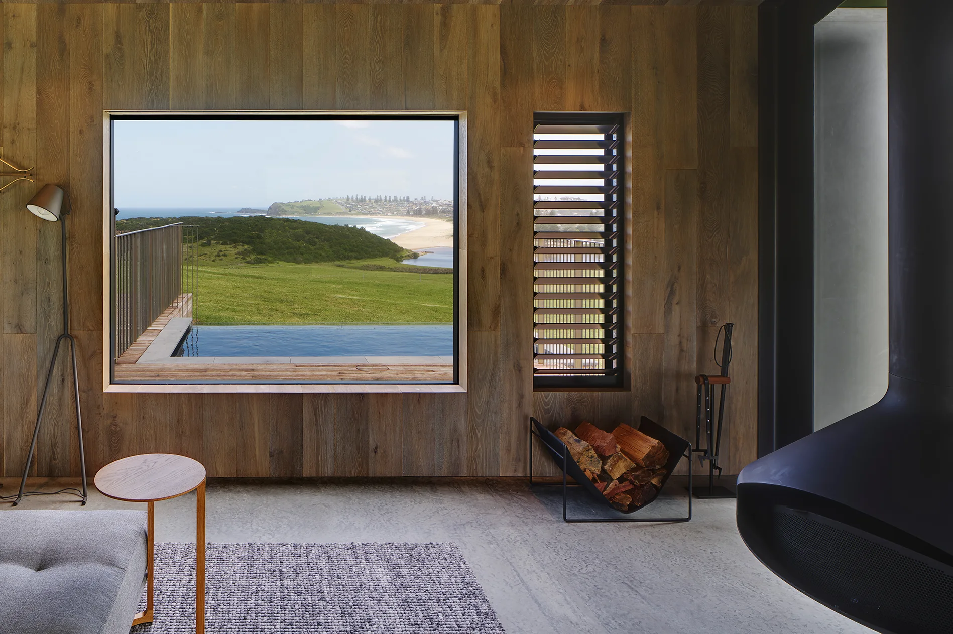 A modern living room features vertical wood paneling, a large picture window framing a coastal view, a concrete floor, and a black fireplace.