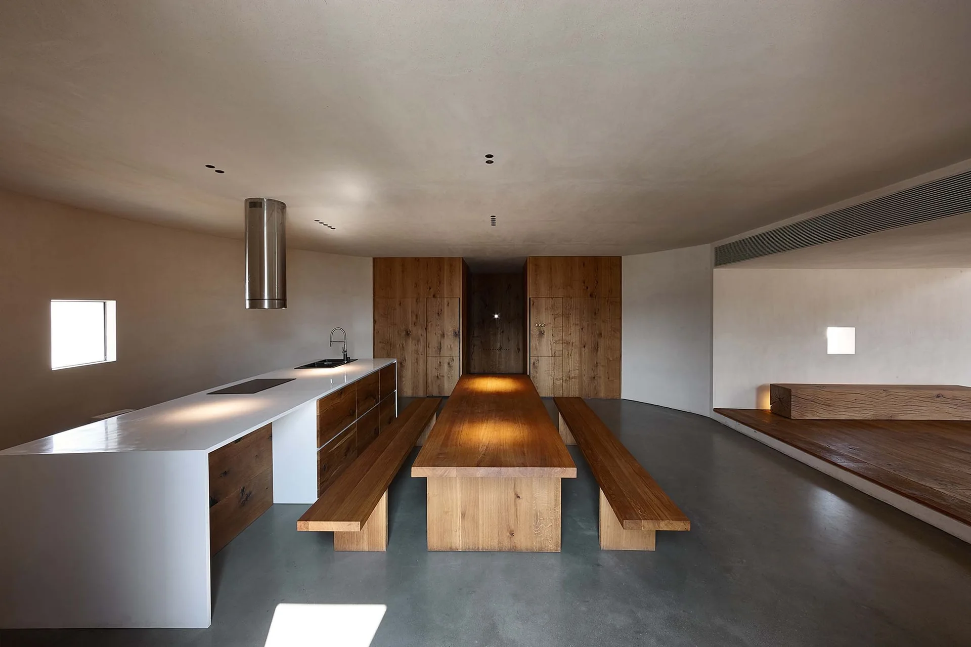 A minimalist open-plan kitchen featuring a long wooden dining table with matching benches, concrete flooring, and a sleek white island.