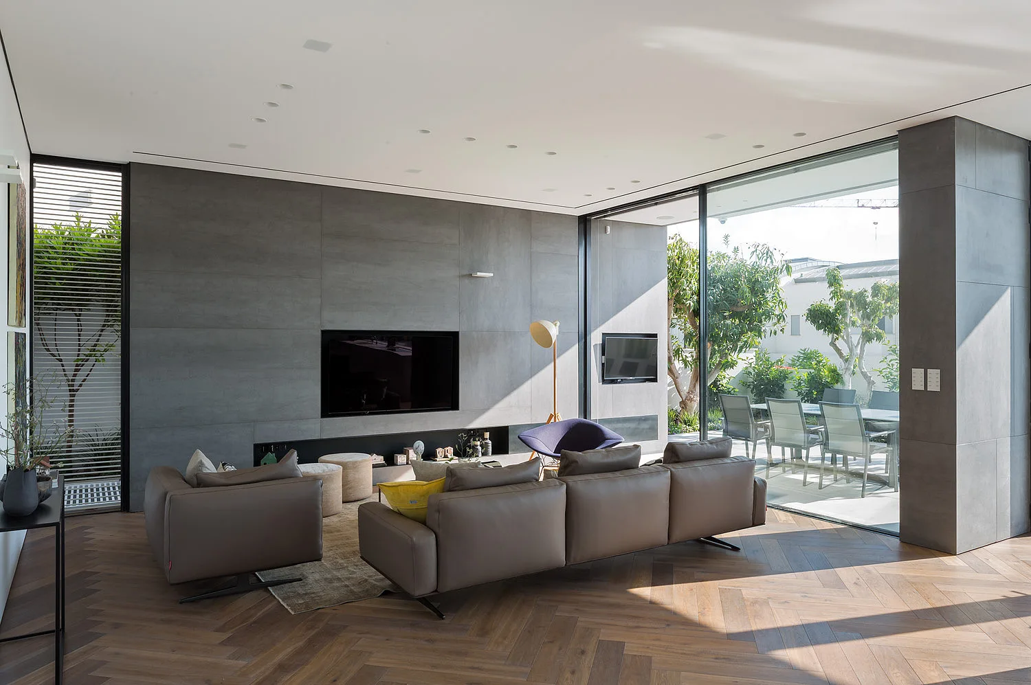 A modern living room featuring a brown leather sectional, a grey paneled media wall, herringbone wood floors, and large sliding glass doors.