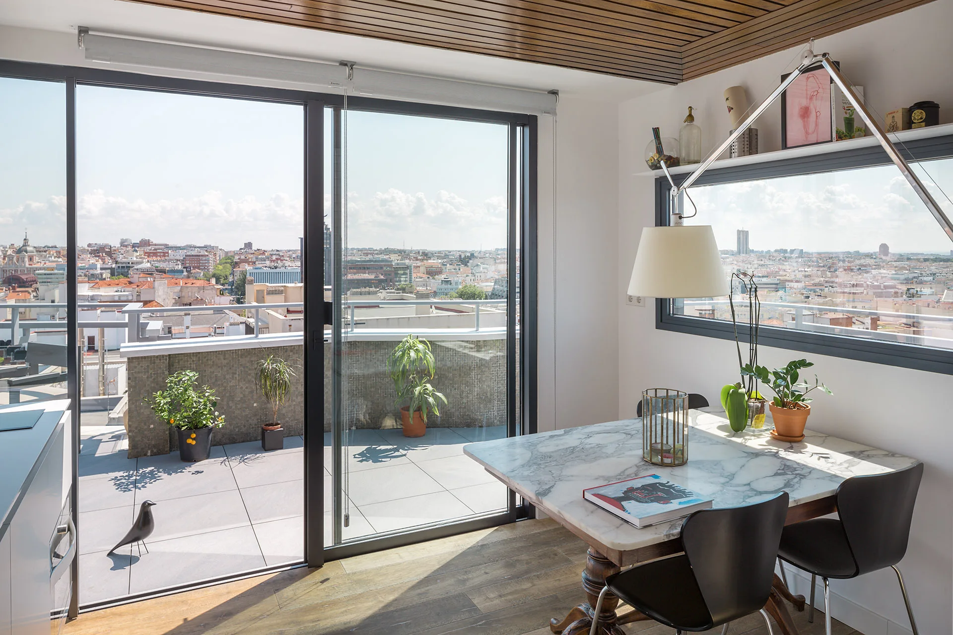 A modern dining room features a marble-topped table and black chairs next to sliding glass doors that open onto a balcony with city views.