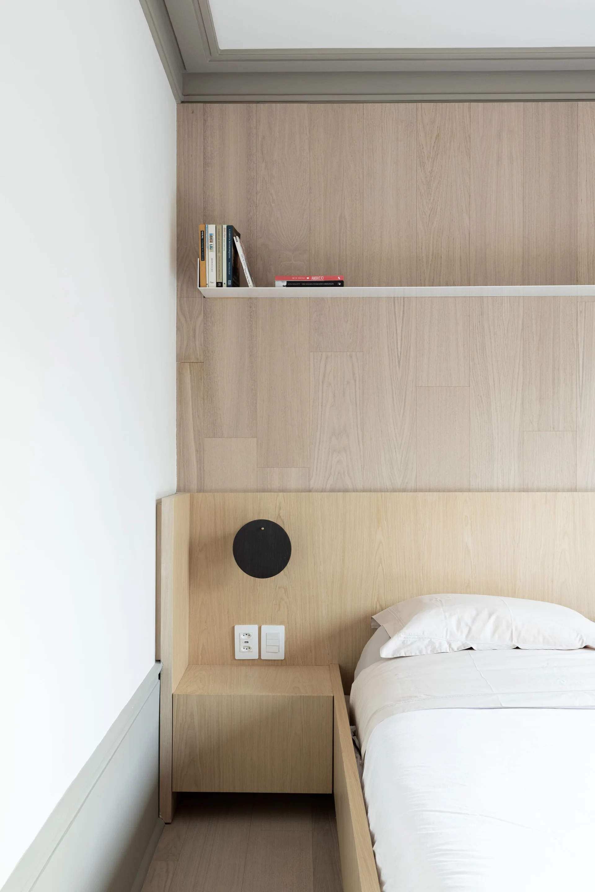 A minimalist bed features a light wood paneled headboard, an integrated nightstand, a circular black sconce, and a thin white floating shelf.