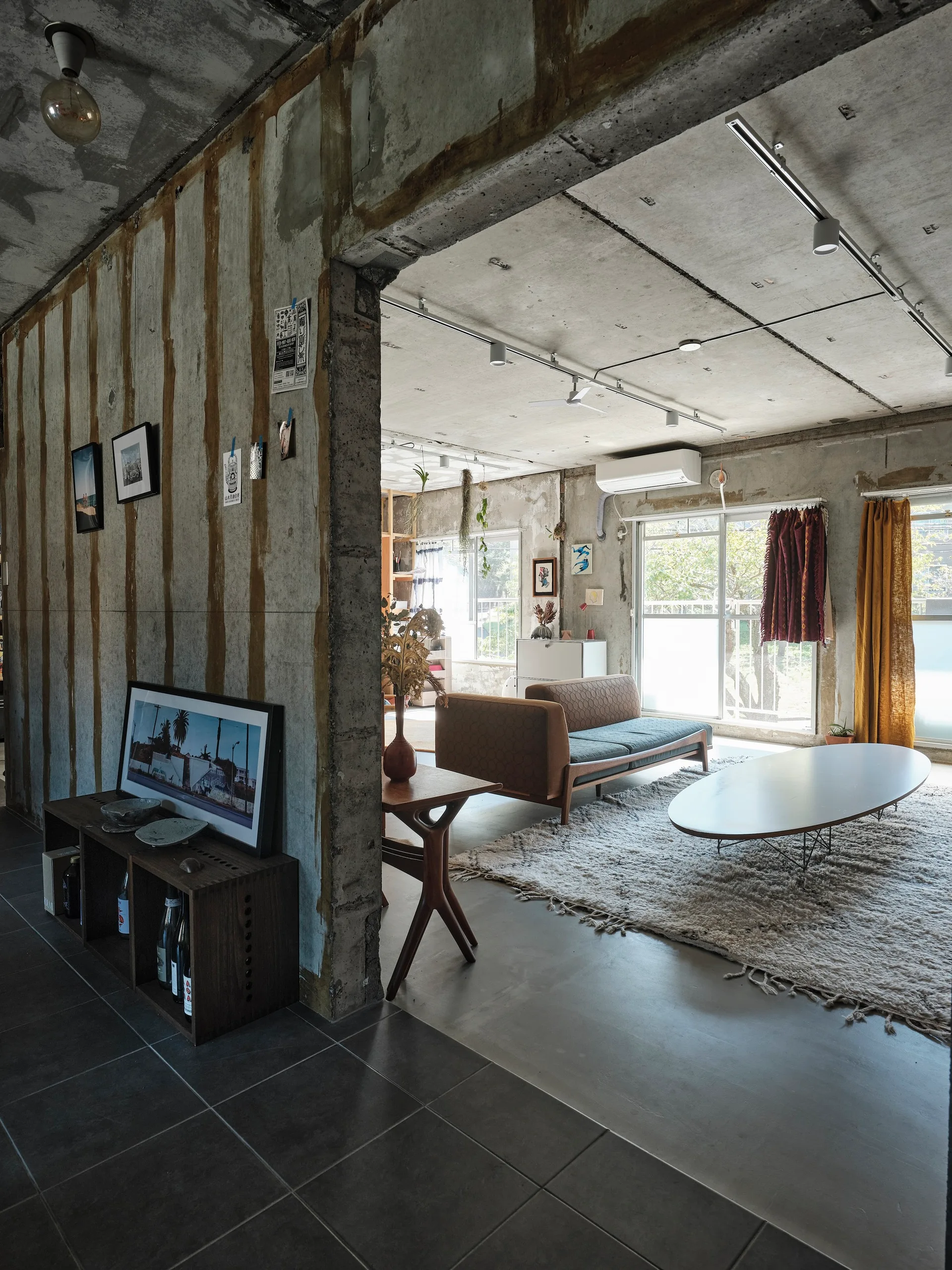 An industrial living room with raw concrete walls, featuring a mid-century modern sofa, an oval coffee table on a thick shag rug, and sliding glass doors.