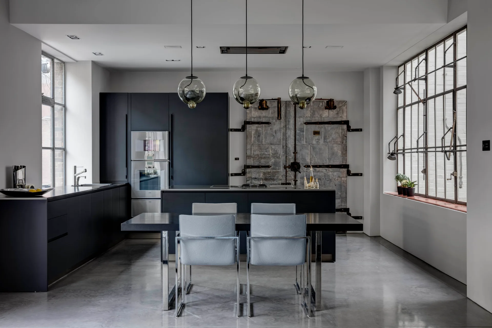 A modern industrial kitchen with charcoal cabinets, a black dining table with gray chairs, polished concrete floors, and glass orb pendant lights.