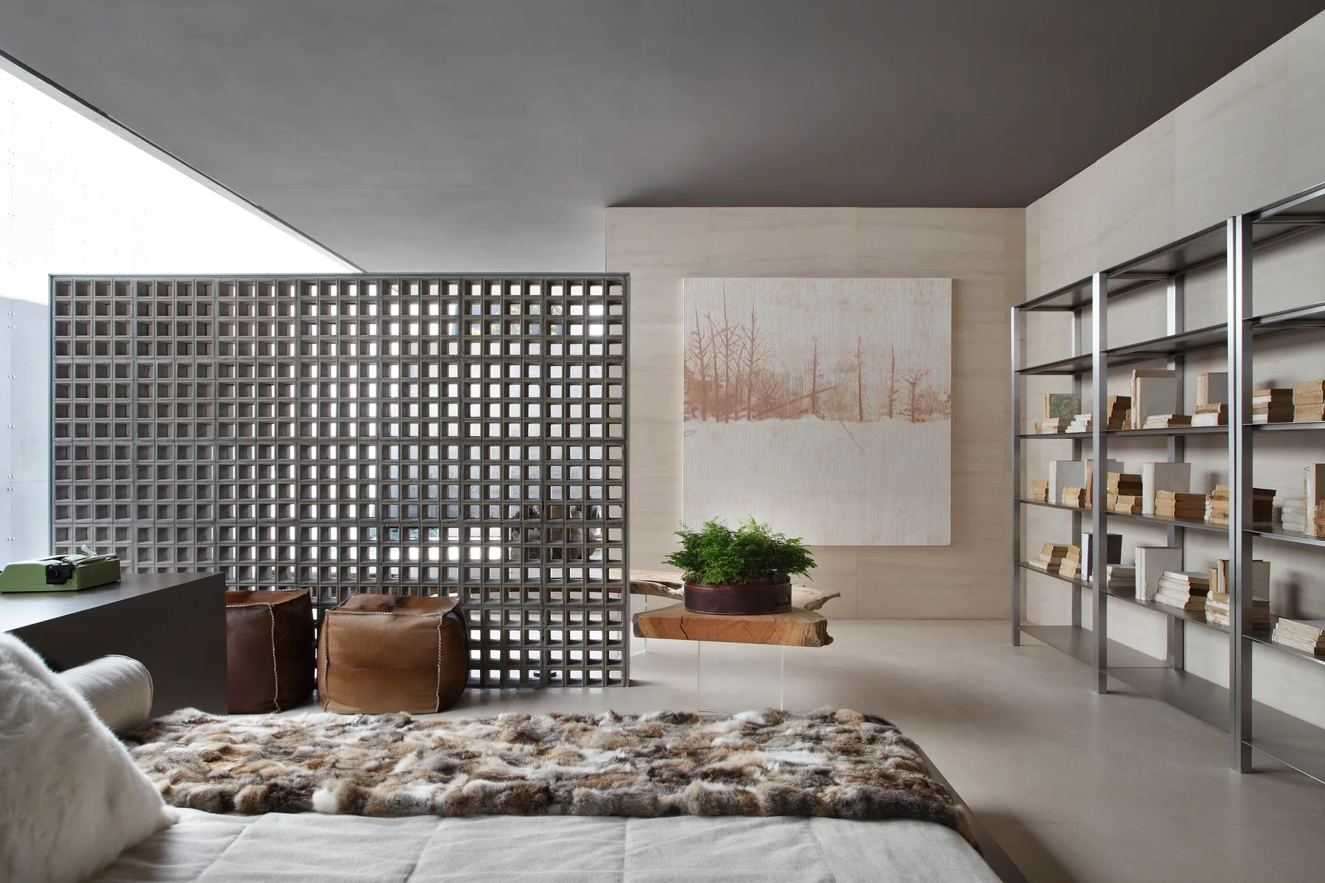 A gray breeze block partition separates a bedroom area from a living space with open metal shelving, a live-edge wooden table, and leather poufs.