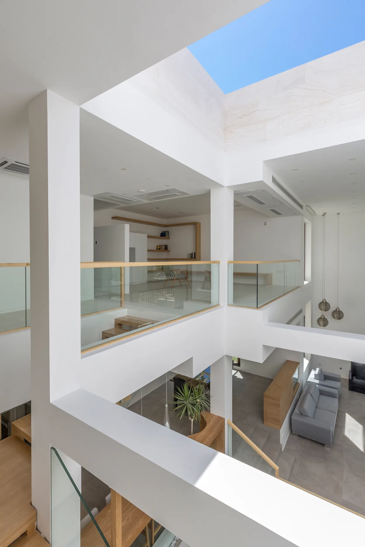 A multi-level interior with white walls, glass railings capped in wood, wooden stairs, and gray sofas on the lower level beneath a large ceiling skylight.