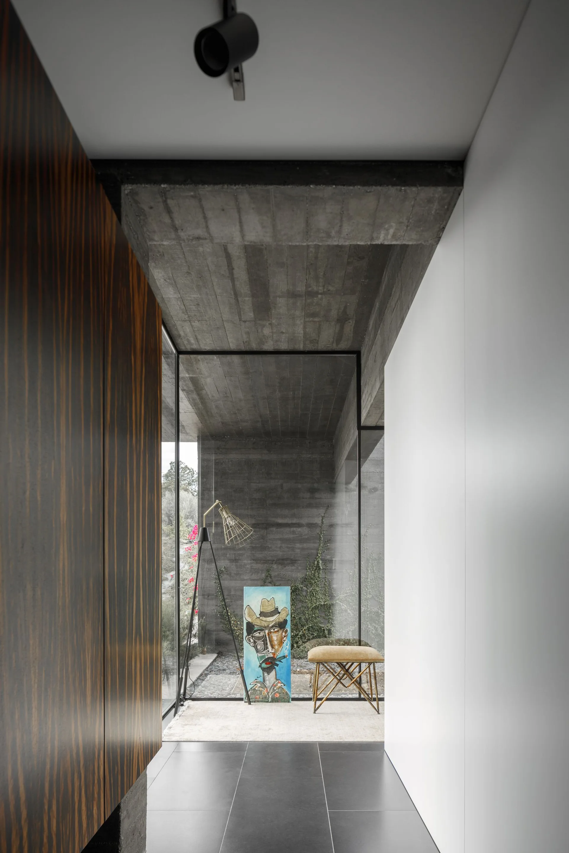 A minimalist hallway featuring raw concrete ceilings, dark wood paneling, a large glass wall, and a decor vignette with a bench and artwork.