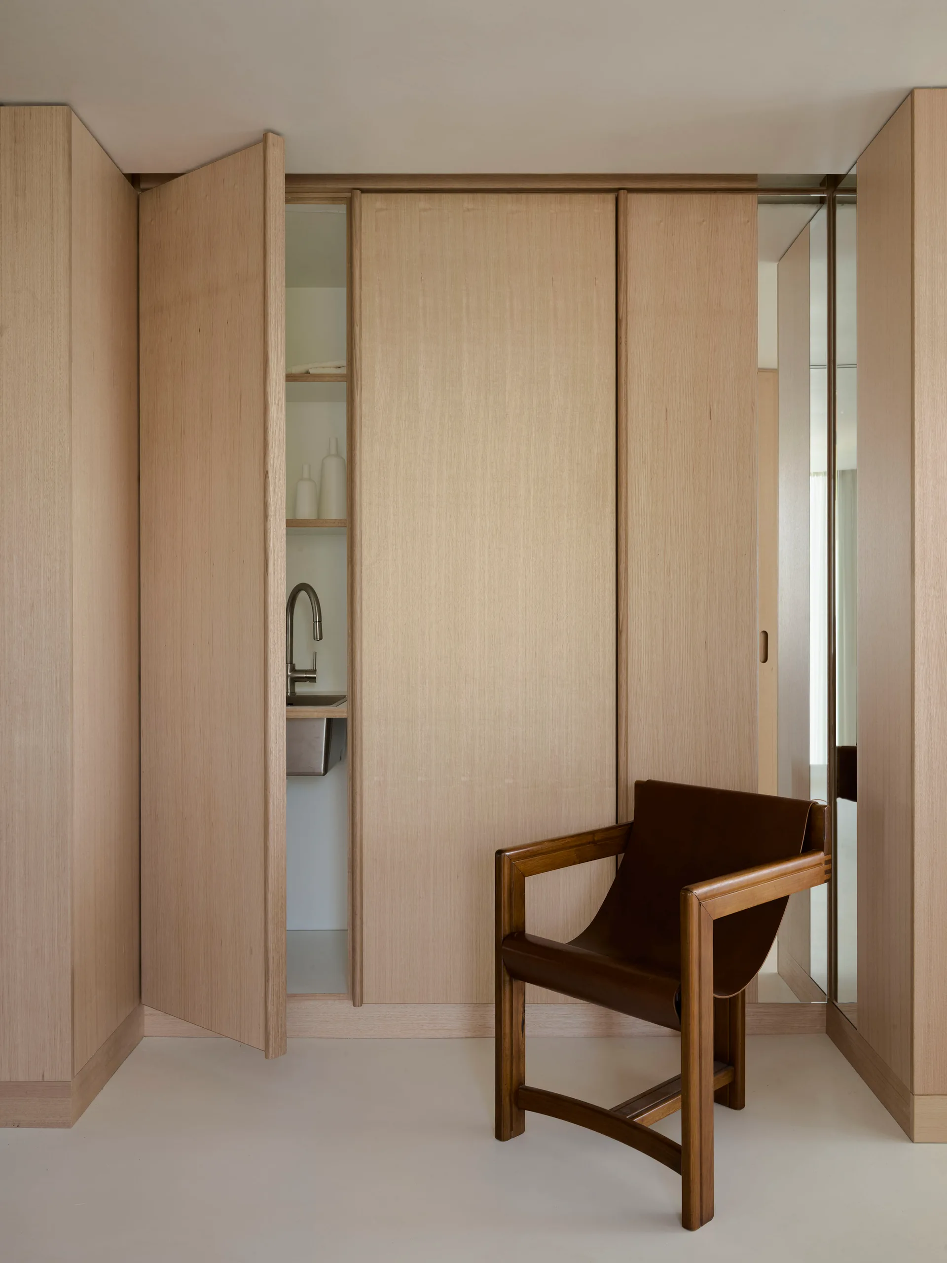 Minimalist light wood cabinetry features an open door revealing a small sink, positioned next to a brown leather armchair.