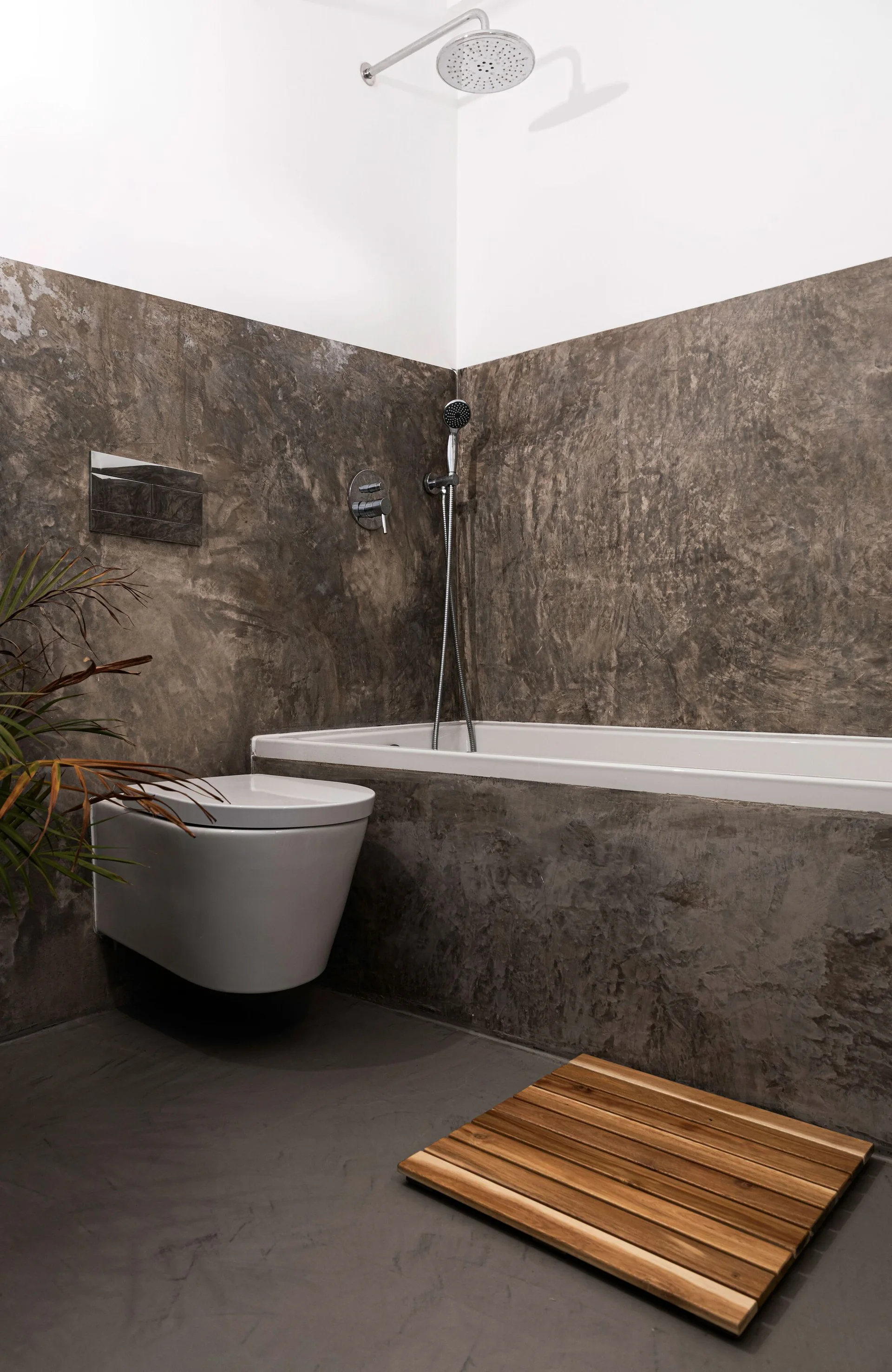 A modern bathroom features textured gray walls, a built-in bathtub, a wall-mounted toilet, and a slatted wooden bath mat.
