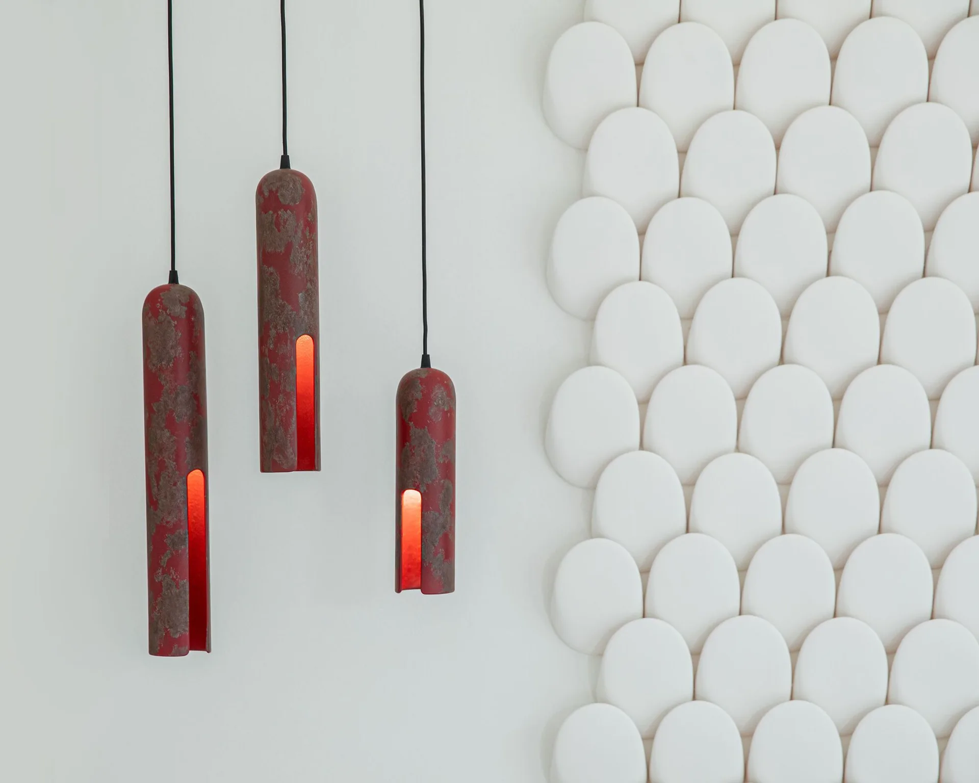 Three distressed red cylindrical pendant lights hang at staggered heights next to a textured white wall featuring a repeating pattern of oval shapes.