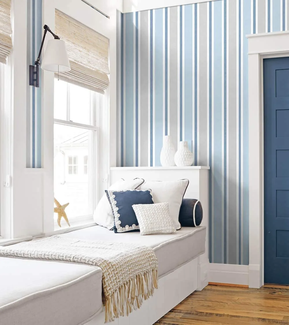 A cozy window seat nook with blue and white striped wallpaper, a cushioned bench, a woven roman shade, and a dark blue door.